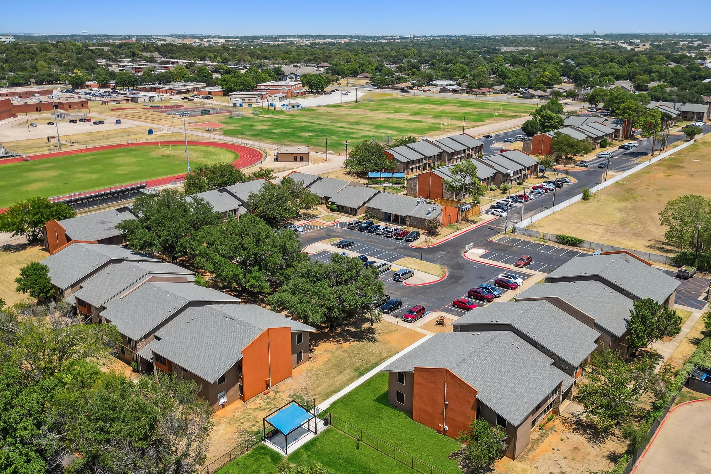 Aerial view of a residential complex featuring several buildings with grey roofs, surrounded by green trees and a parking area. A sports field and track are visible in the background, along with additional structures. The area is sunny and well-maintained, with a clear sky above.