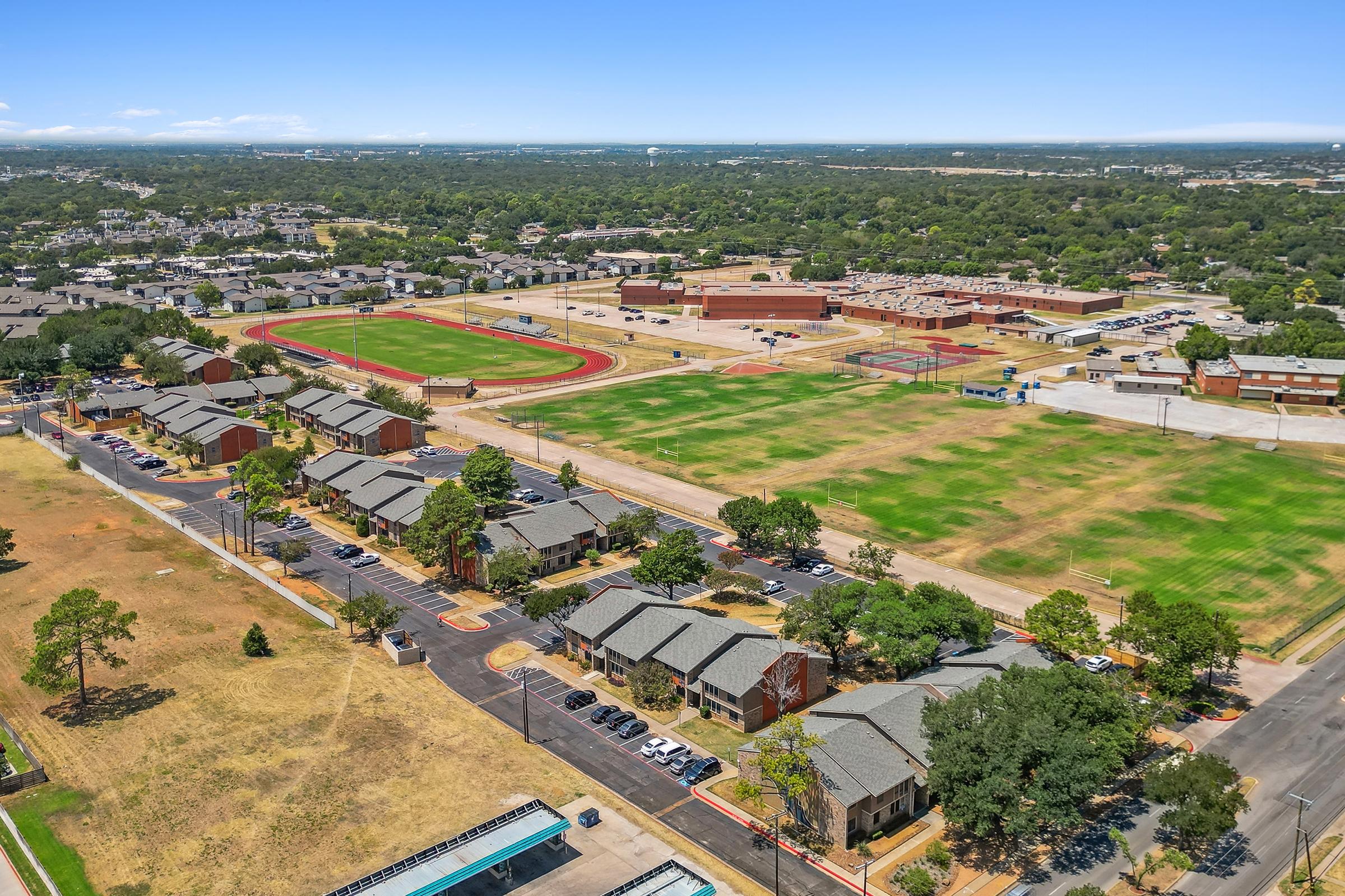Aerial view of a sprawling residential area with multiple buildings lined along a street. In the background, there's a large school complex with sports fields, including a running track and open grassy areas. Surrounding landscapes are green with trees and open spaces, under a clear blue sky.