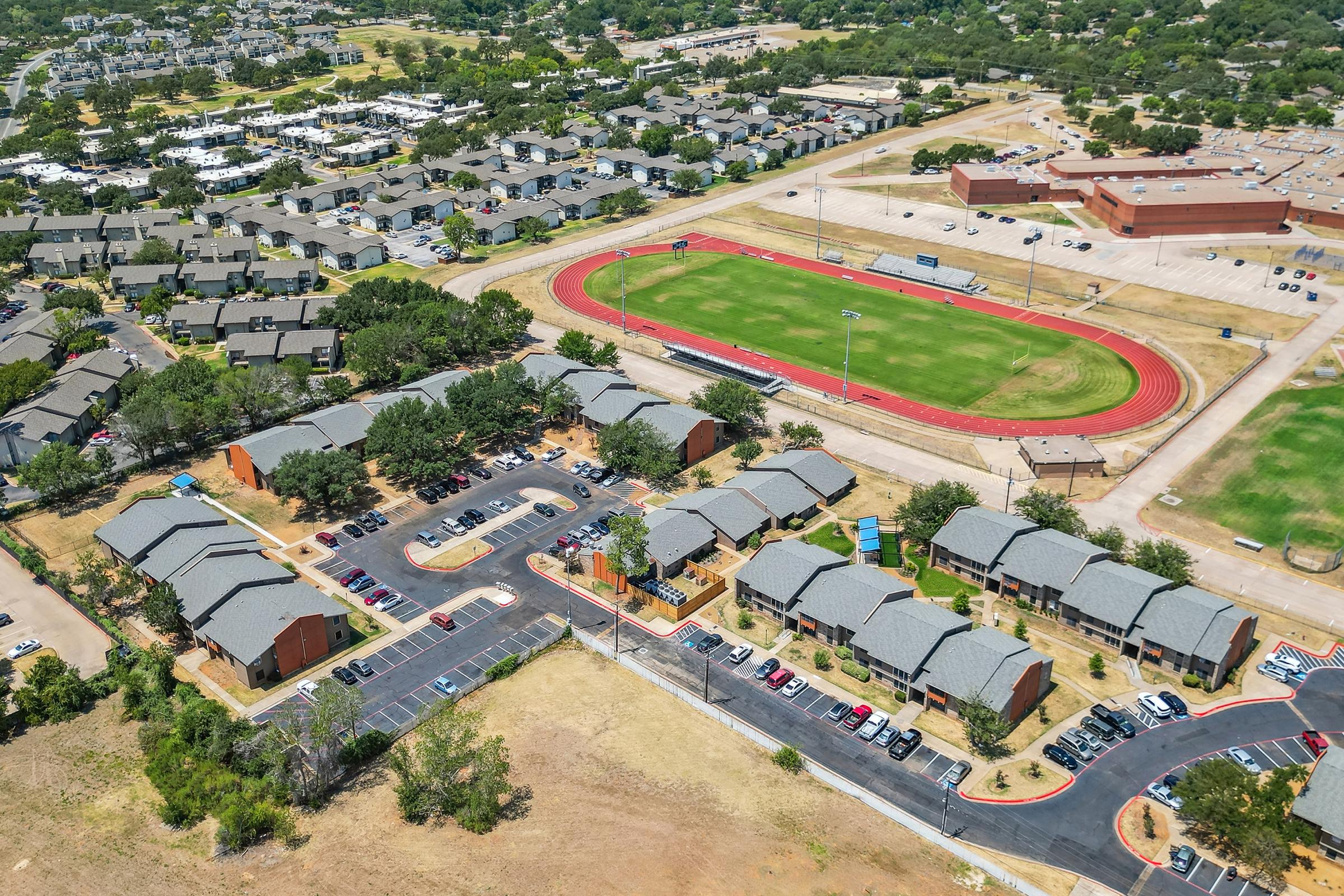 Aerial view of a residential area with low-rise apartments, surrounded by greenery. A red running track and field are visible nearby, along with a larger building that appears to be a school. The landscape includes parking lots and open grassy areas.