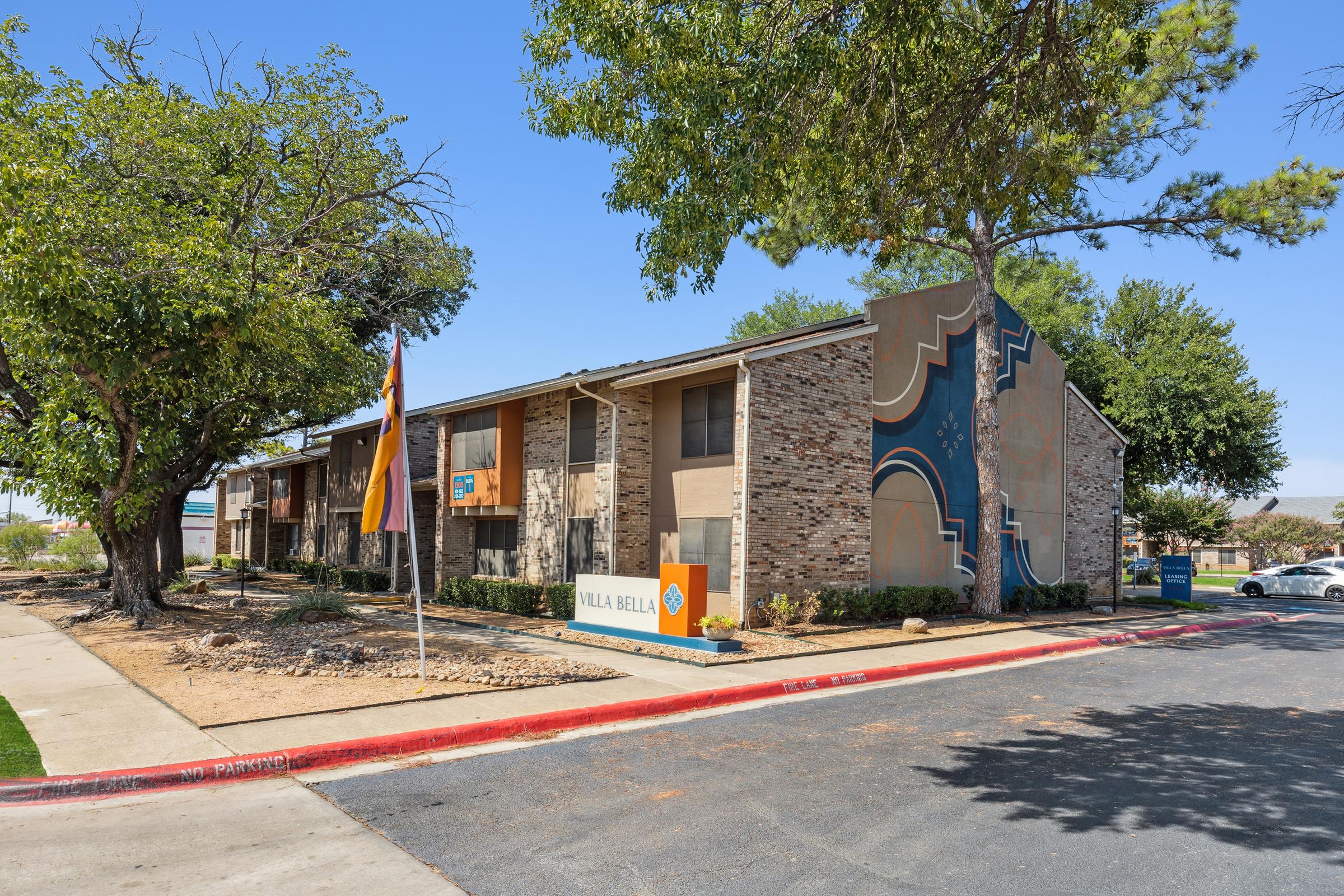 Apartment complex named Villa Bella with a distinctive mural on the side. The building is surrounded by trees and landscaping, and a flag is visible in front. There is a parking lot nearby, and the area appears well-maintained.