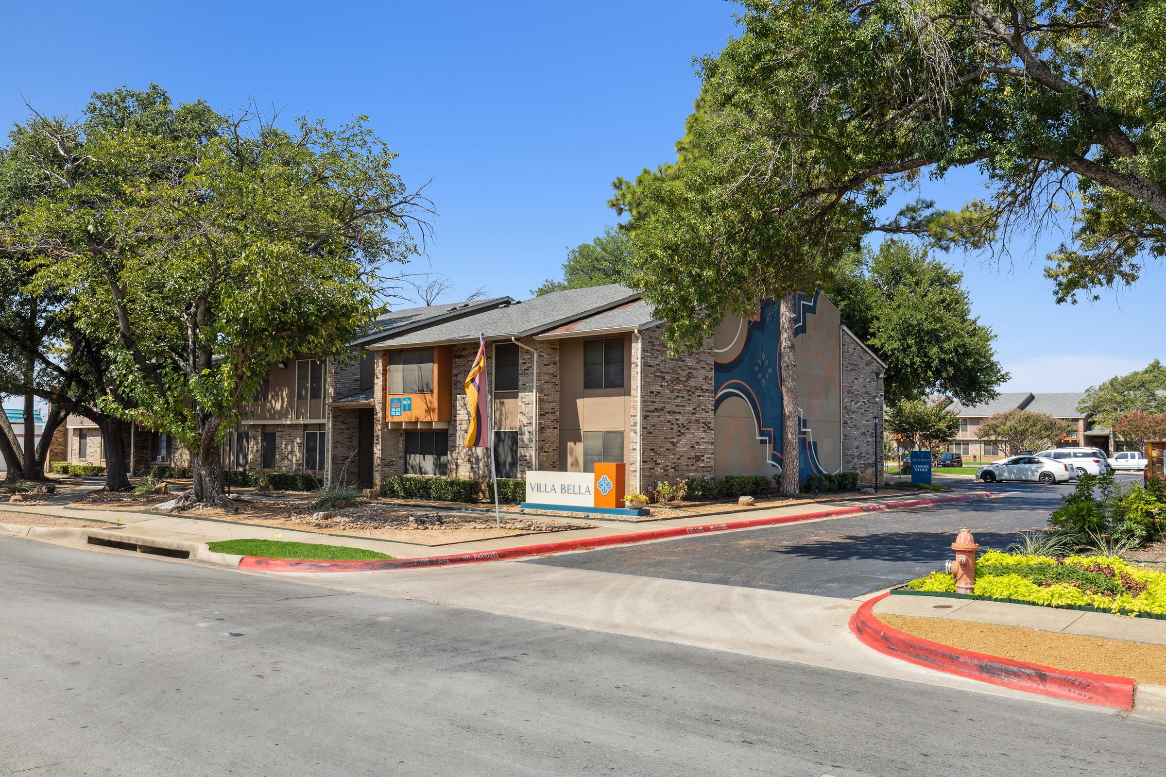 Exterior view of a residential complex named "Villa Bella," featuring multiple buildings, landscaped areas, and clear blue skies. The parking lot is visible with a few cars parked, and trees provide shade around the property. A colorful mural is displayed on one of the buildings.