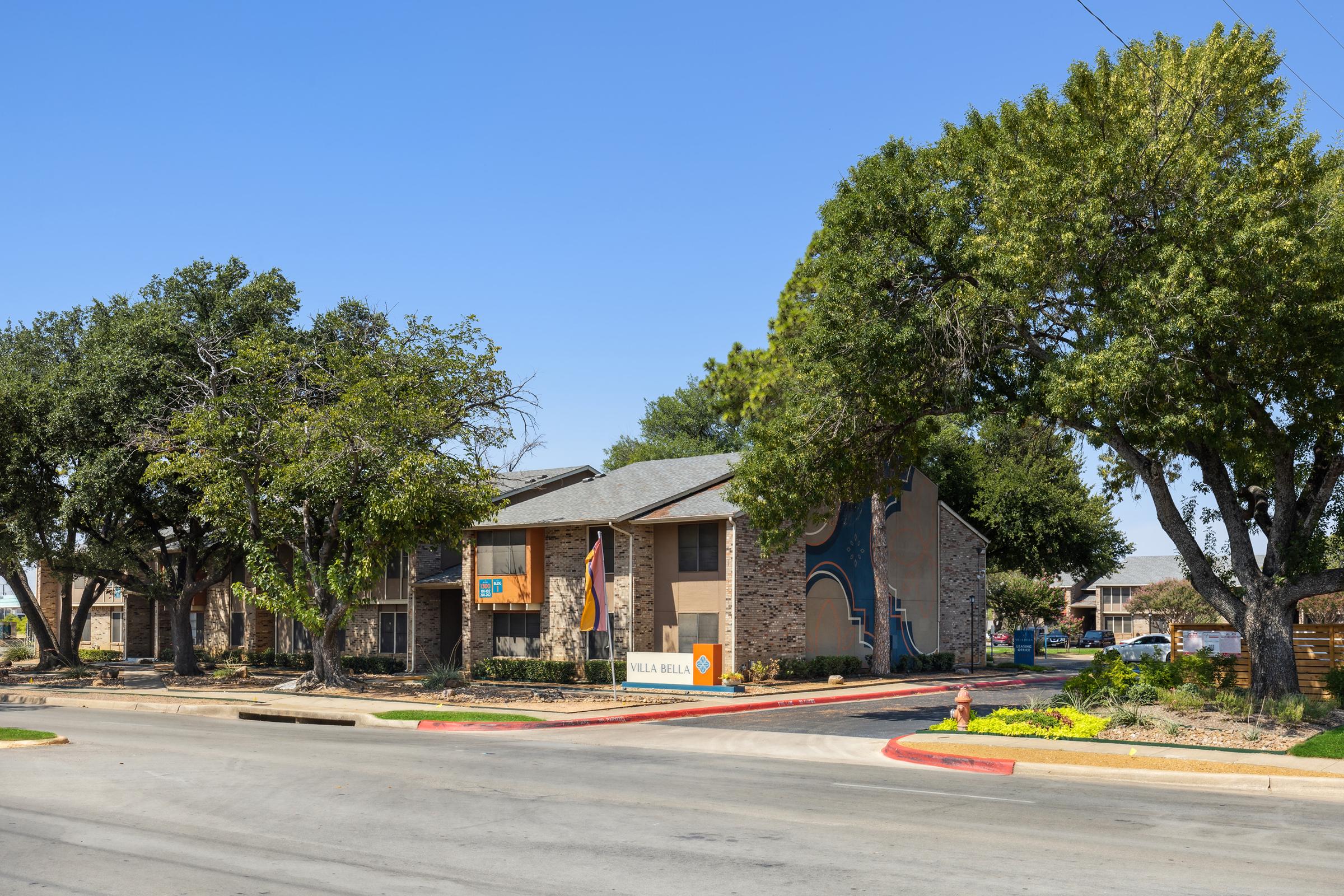 A view of a residential apartment complex with a sign that reads "Villa Bella." The building features a mixture of stone and wooden elements, surrounded by large trees and landscaping. The street in front is paved, with a clear blue sky overhead.