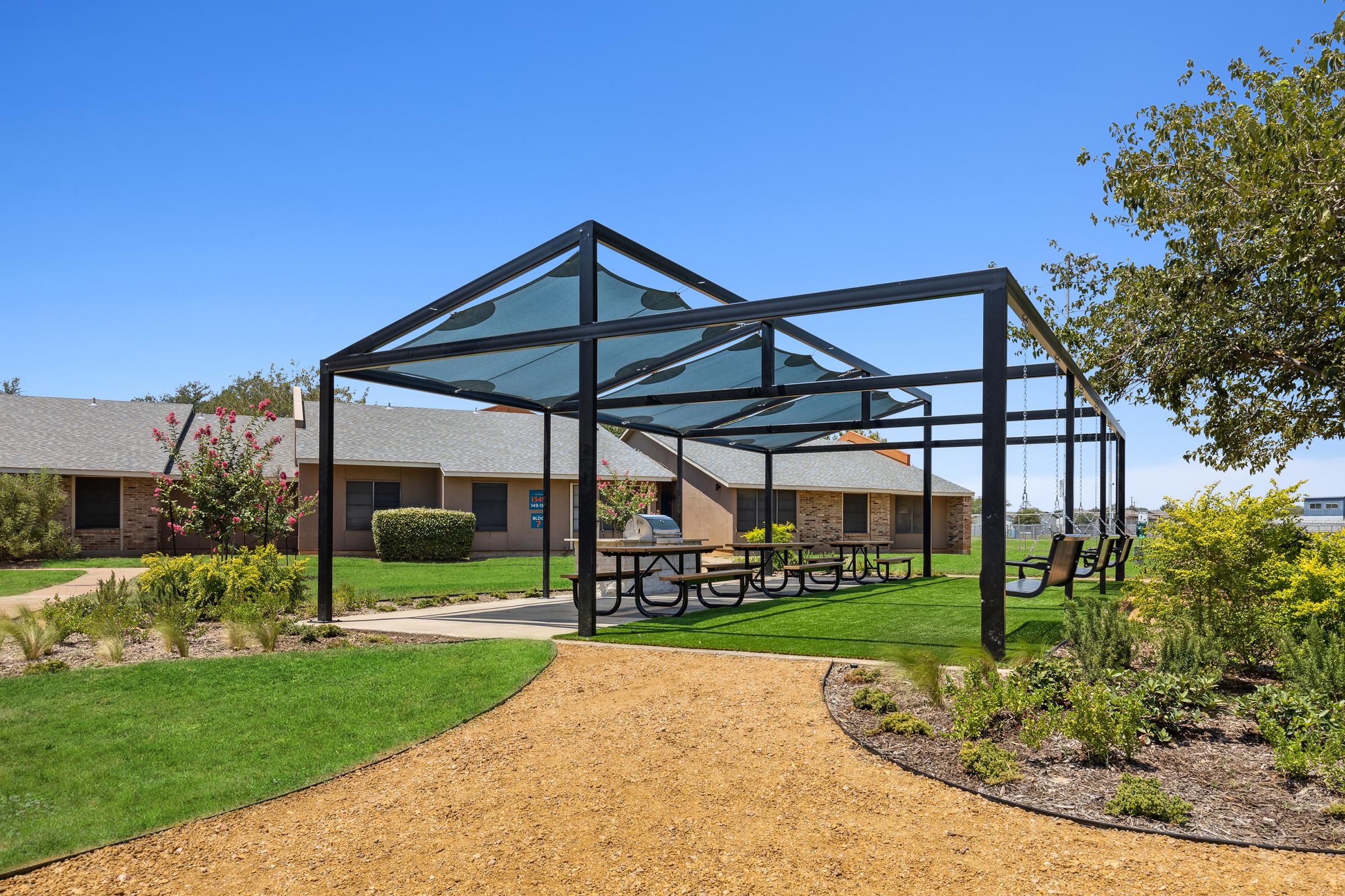 A modern outdoor pavilion with a shade structure featuring picnic tables, surrounded by well-maintained grass and landscaping. In the background, there are buildings with a clear blue sky above. The scene suggests a pleasant and inviting space for gatherings or relaxation.