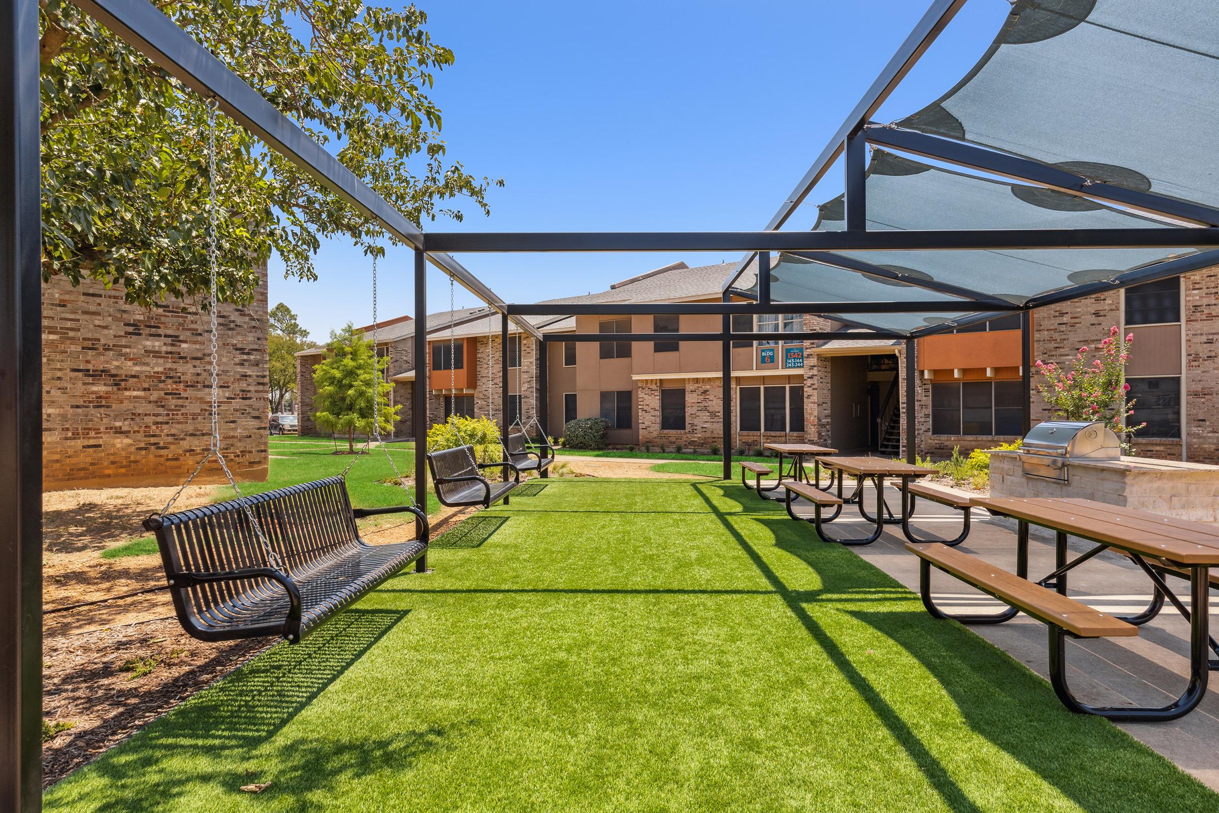 A green outdoor space featuring shaded seating areas with black benches and wooden picnic tables. The area is surrounded by buildings, and there is a grassy lawn under the shade structures. The sky is clear and blue, creating a bright and inviting atmosphere.