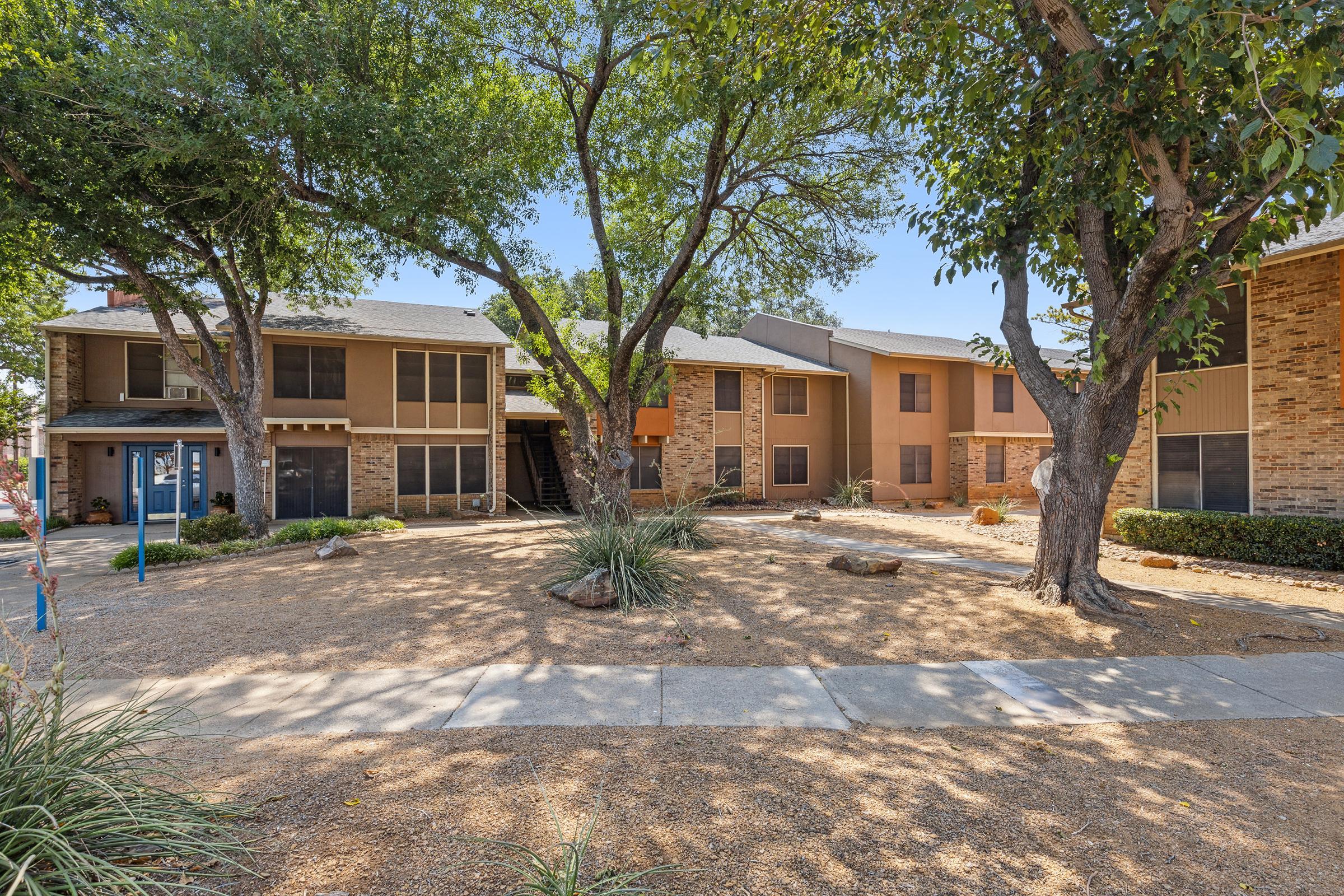 A multi-unit residential building featuring a combination of stone and siding exteriors, surrounded by trees and shrubs. A sandy, open area with landscaped plants and a pathway leads to the entrance. The scene is bright and inviting, showcasing a well-maintained outdoor space.