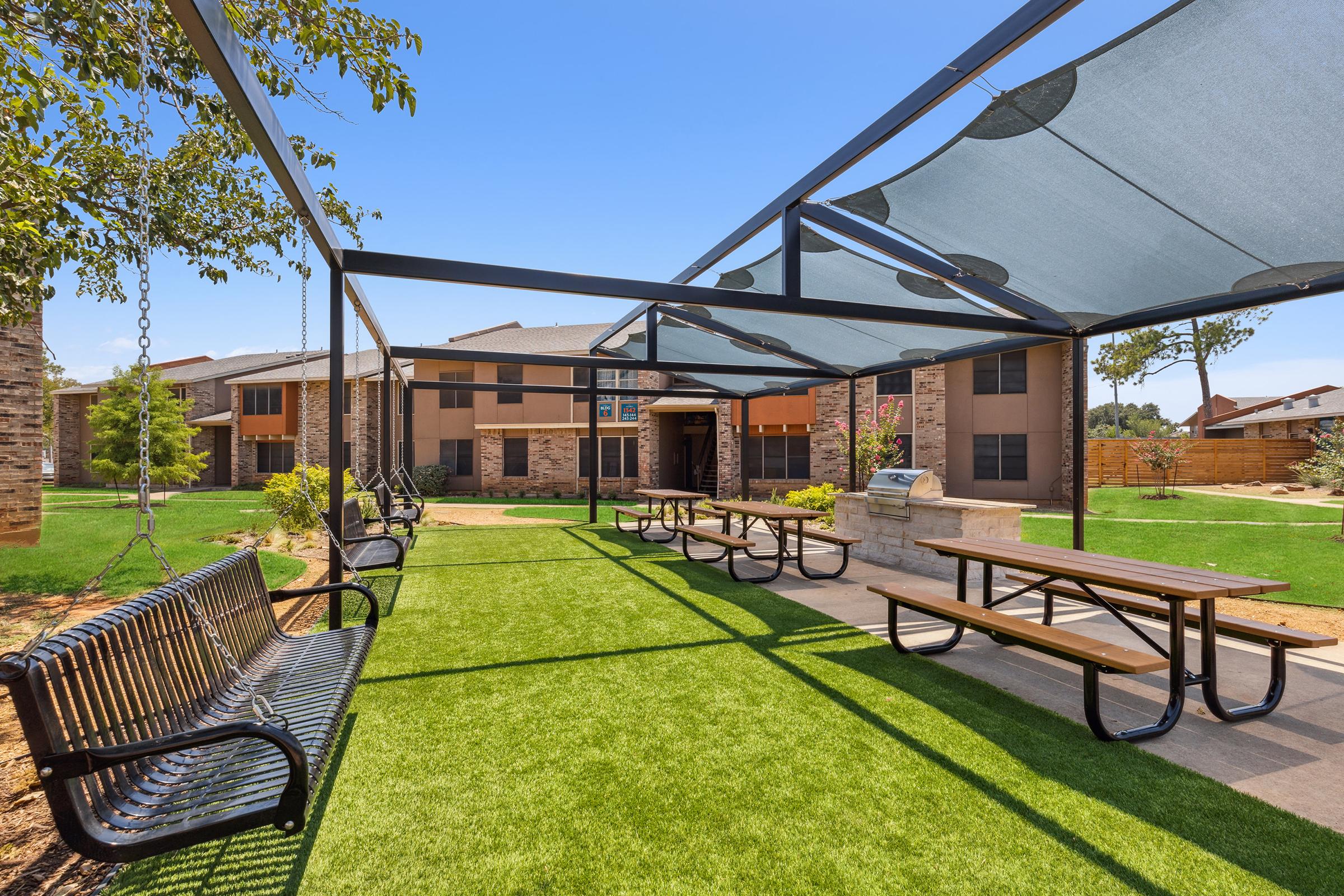 Outdoor recreational area featuring shaded seating with benches and picnic tables on artificial grass, surrounded by landscaped greenery and residential buildings in the background.