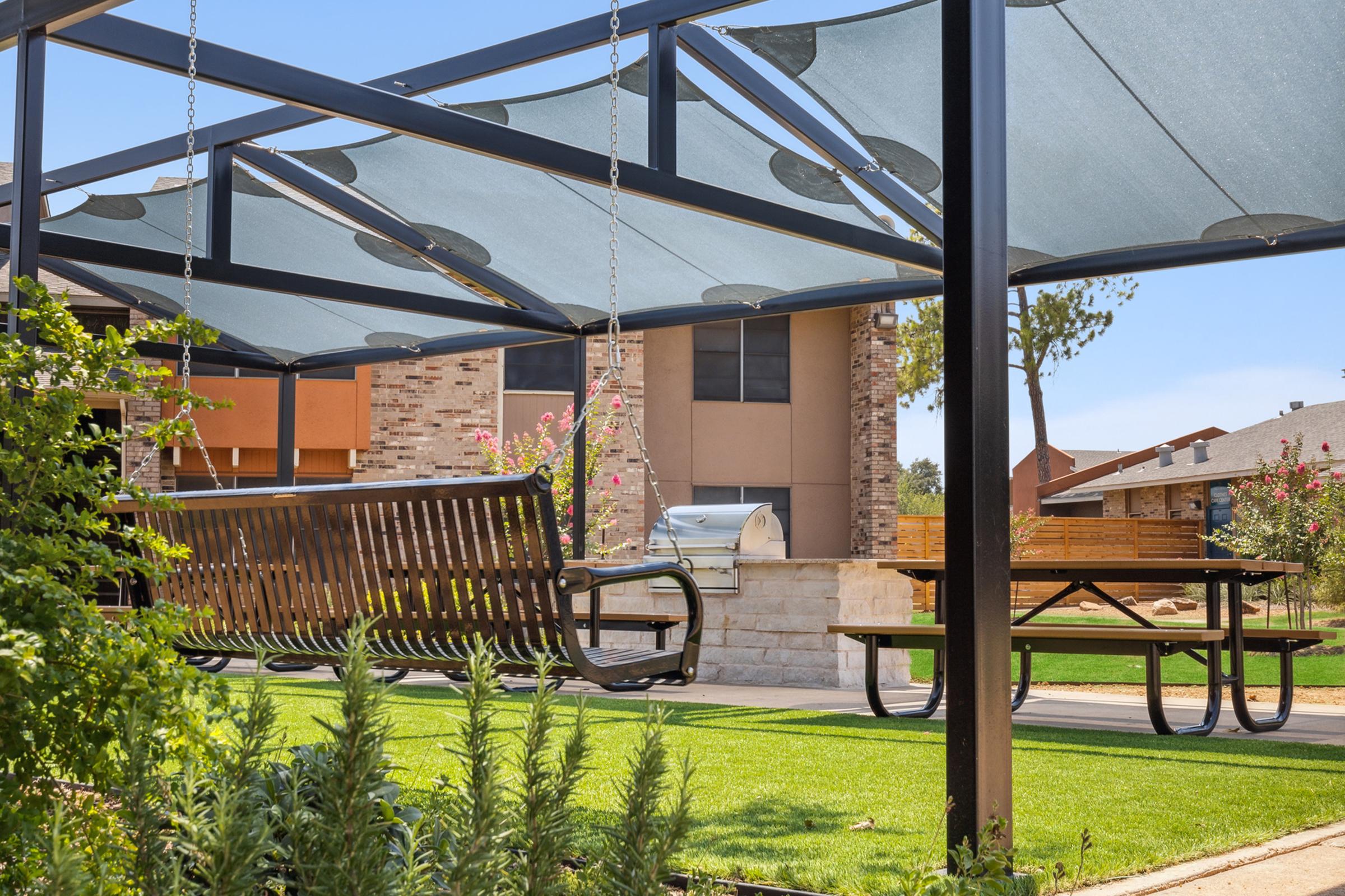 A shaded outdoor area featuring a wooden swing and a picnic table on a grassy lawn. In the background, there is a building with a stone exterior and a grill nearby. The space is surrounded by plants and trees, creating a pleasant and inviting environment.