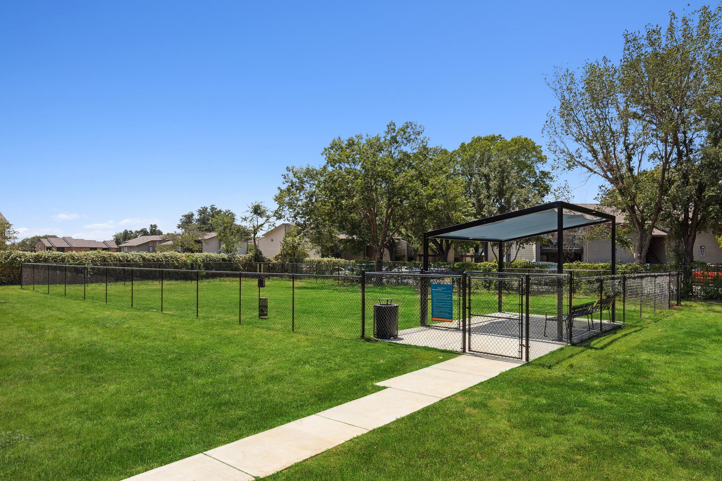 A sunny park area featuring a spacious, fenced dog park with a shaded seating area. Well-maintained grass surrounds the park, and there are trees lining the background. A pathway leads into the park, with residential buildings visible in the distance under a clear blue sky.