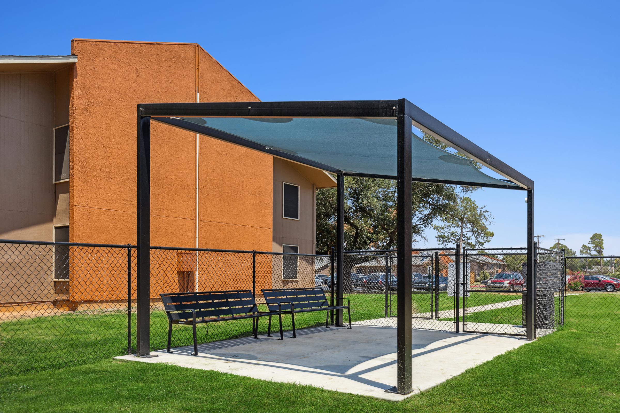 A shaded picnic area with benches is surrounded by a black chain-link fence, set on a green lawn. In the background, there are orange and beige apartment buildings under a clear blue sky.