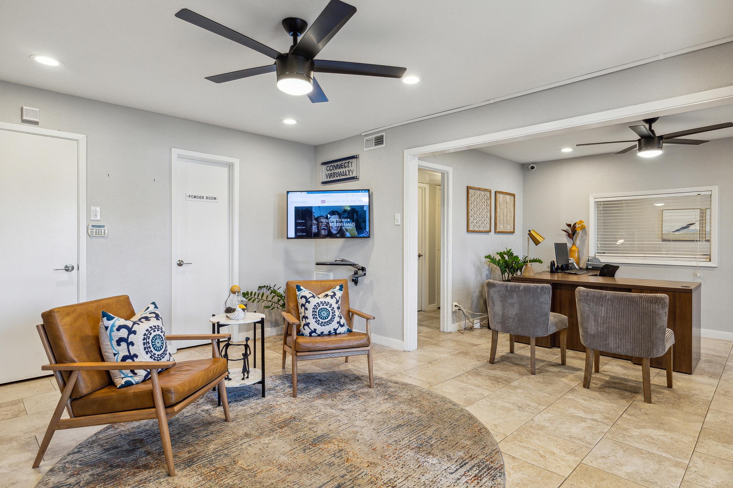 A modern waiting area featuring two brown armchairs with blue patterned pillows, a circular area rug, a small table, and a wall-mounted TV displaying a website. There is a desk with two gray chairs, a few plants, and light gray walls with decorative artwork. Ceiling fans provide ventilation.