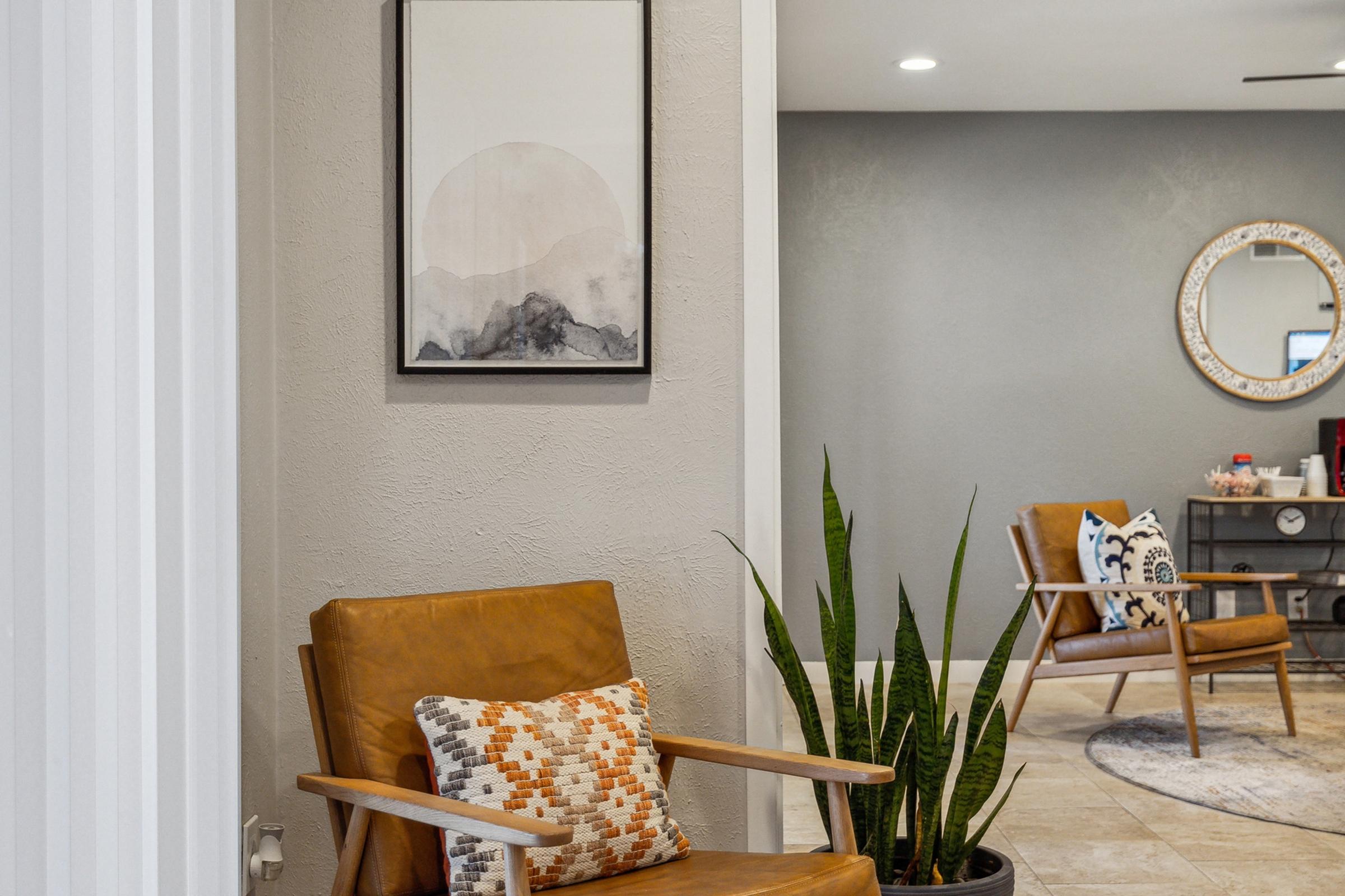A cozy interior space featuring a brown leather armchair with a patterned pillow, a minimalistic wall art piece depicting a faded landscape, and a tall indoor plant. In the background, another seating area can be seen with a round mirror and stylish decor elements.