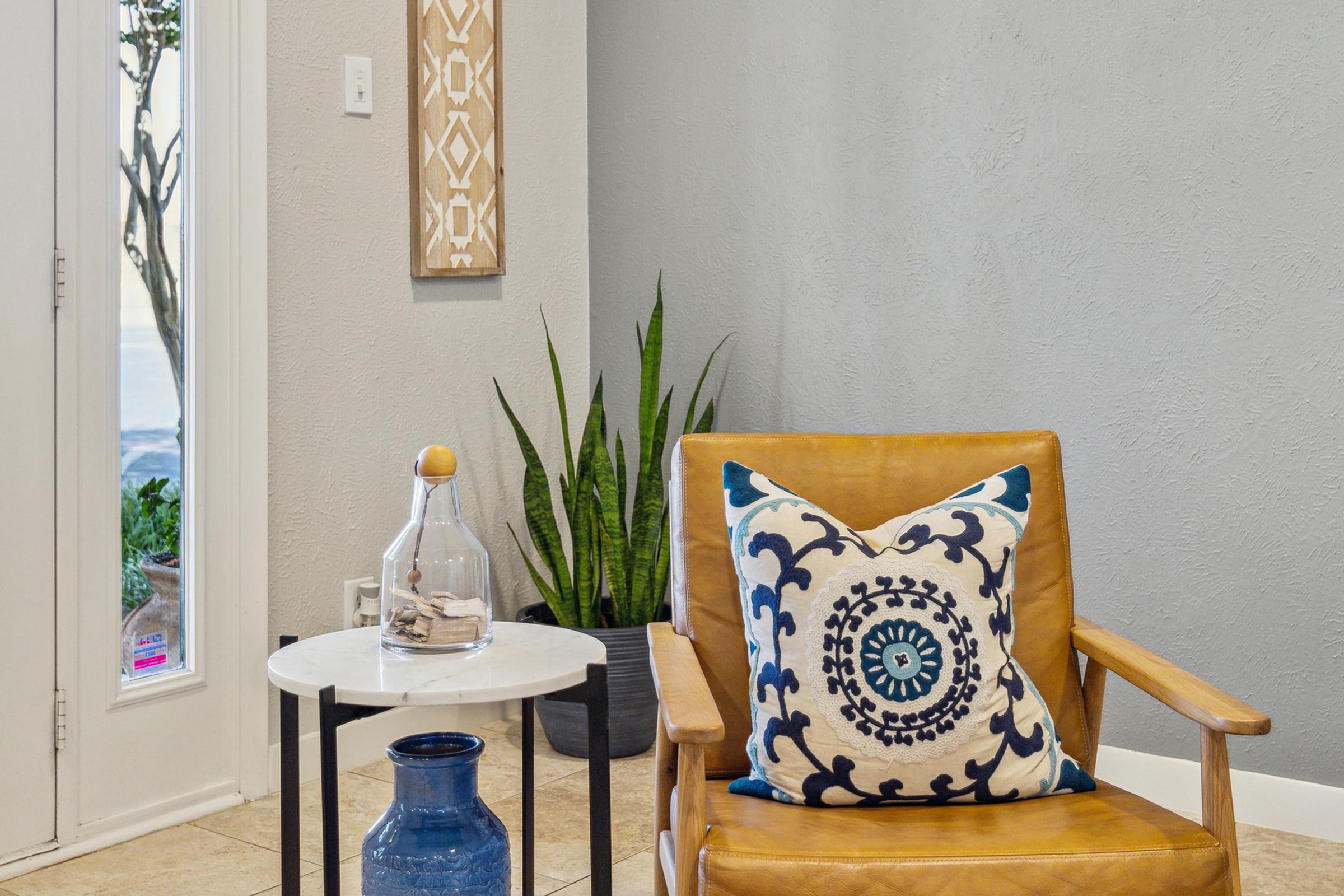A cozy living space featuring a stylish yellow armchair with a decorative blue and white pillow. Next to it, a small white marble side table holds a glass bottle and a blue vase. A tall green plant adds a touch of nature, and a textured wall art piece hangs nearby, enhancing the modern ambiance.