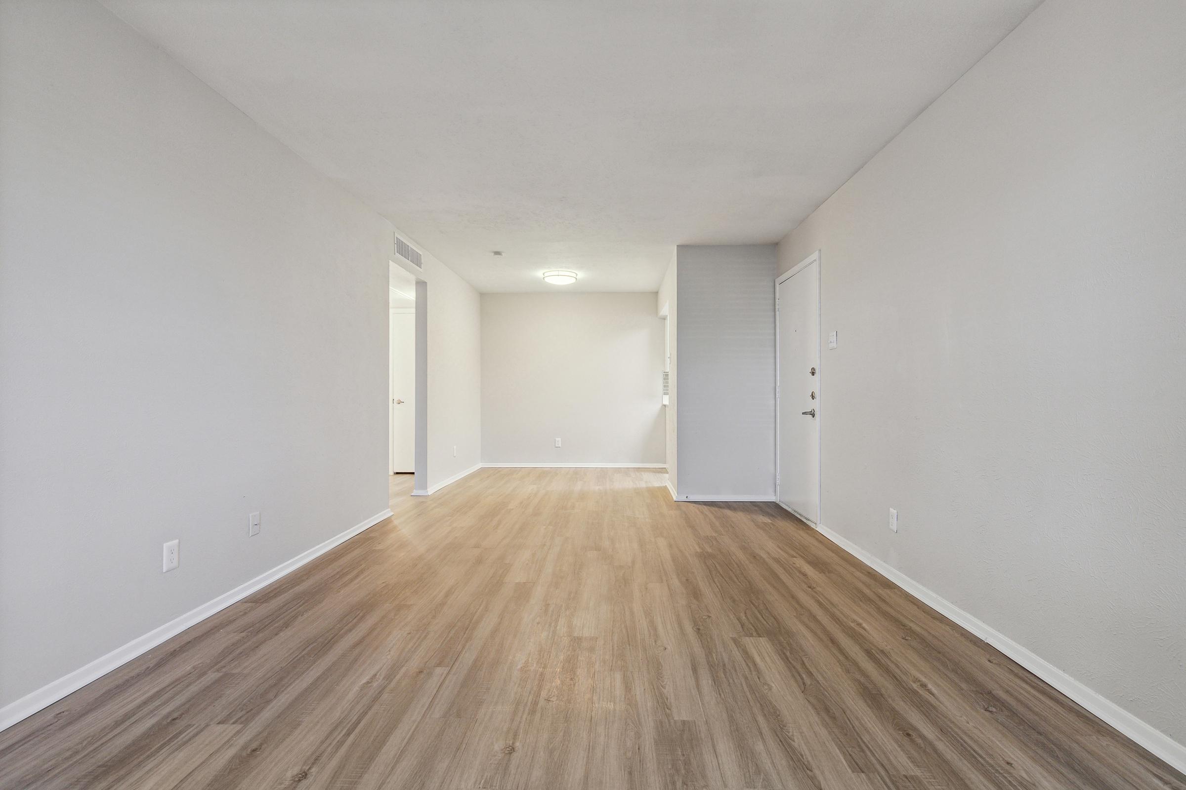 A spacious, empty room with light wood flooring and neutral-colored walls. The far end features a doorway leading to another room, while the left side shows an entrance door. Natural light brightens the space, creating a welcoming and airy atmosphere.