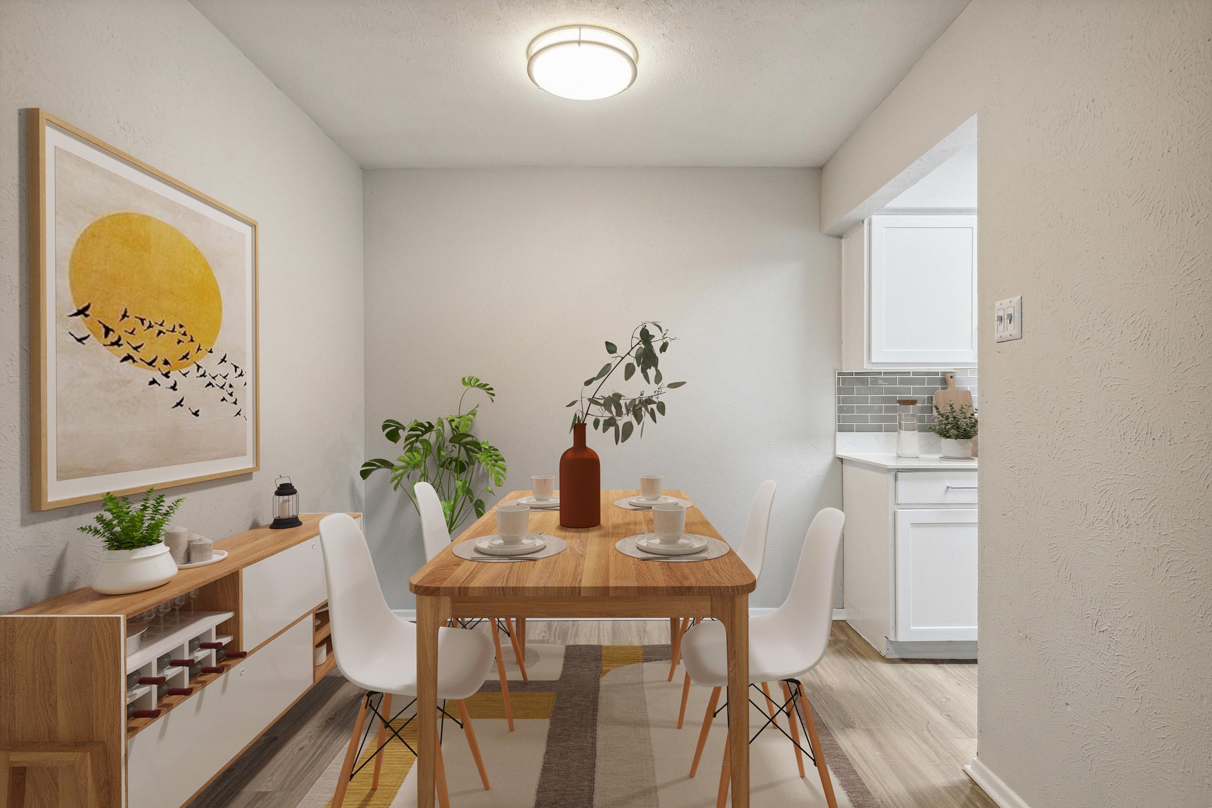 A cozy dining area featuring a wooden table set for two, white chairs, and a minimalist decor. A large artwork with a sun and birds hangs on the wall. A green plant adds a touch of nature, while a sideboard displays decorative items. The kitchen is visible in the background with white cabinetry and a modern design.