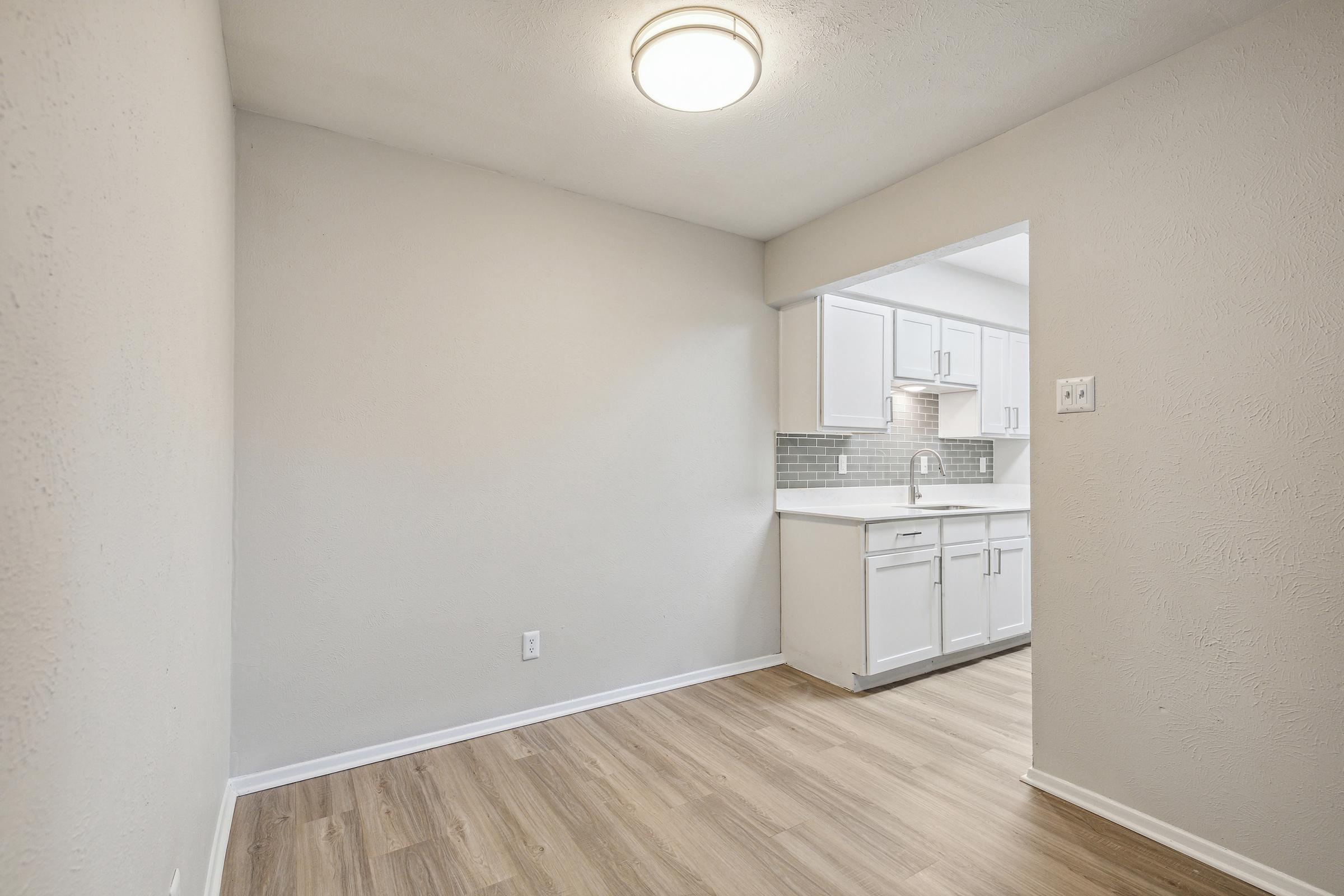 A well-lit, modern kitchen with white cabinetry and a gray backsplash. The space features a light-colored wall, laminate flooring, and a circular ceiling light, creating a clean and inviting atmosphere.