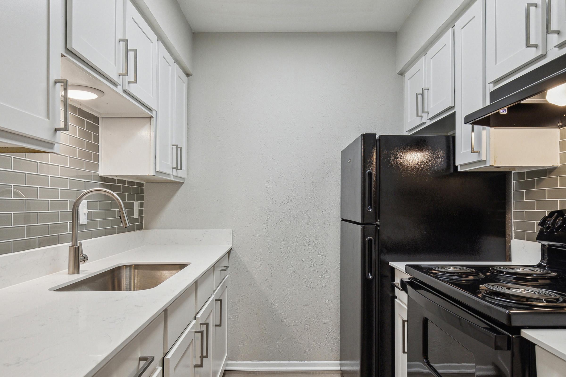 Modern kitchen featuring white cabinetry with silver hardware, a stainless steel sink, and a black refrigerator. The countertop is white with a marble-like finish. The backsplash consists of gray tiles, and the kitchen includes a black stove with an oven. The space is well-lit and has a clean, contemporary design.