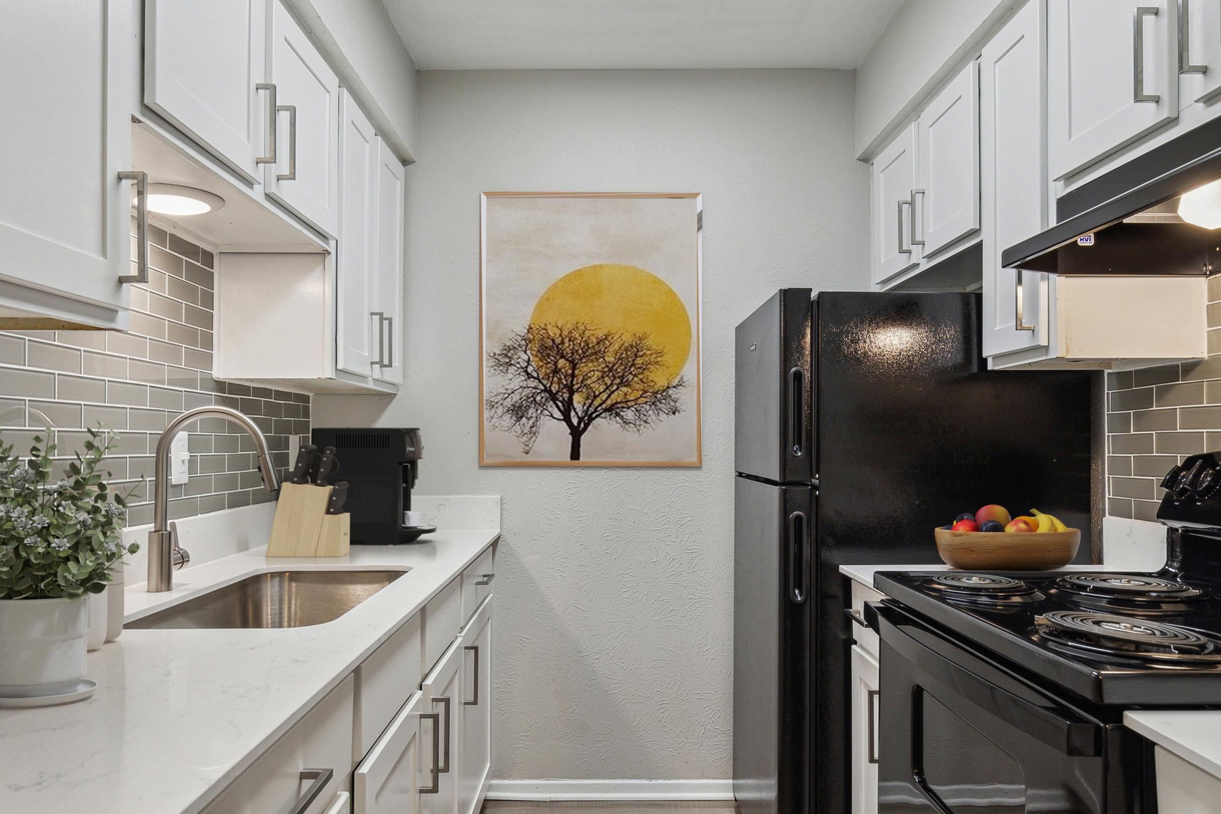 A modern kitchen featuring white cabinetry, a stainless steel sink, and black appliances. There is a framed artwork on the wall depicting a tree against a large yellow sun. A bowl of colorful fruit sits on the stove, adding a touch of vibrancy to the space. Greenery is visible on the countertop.