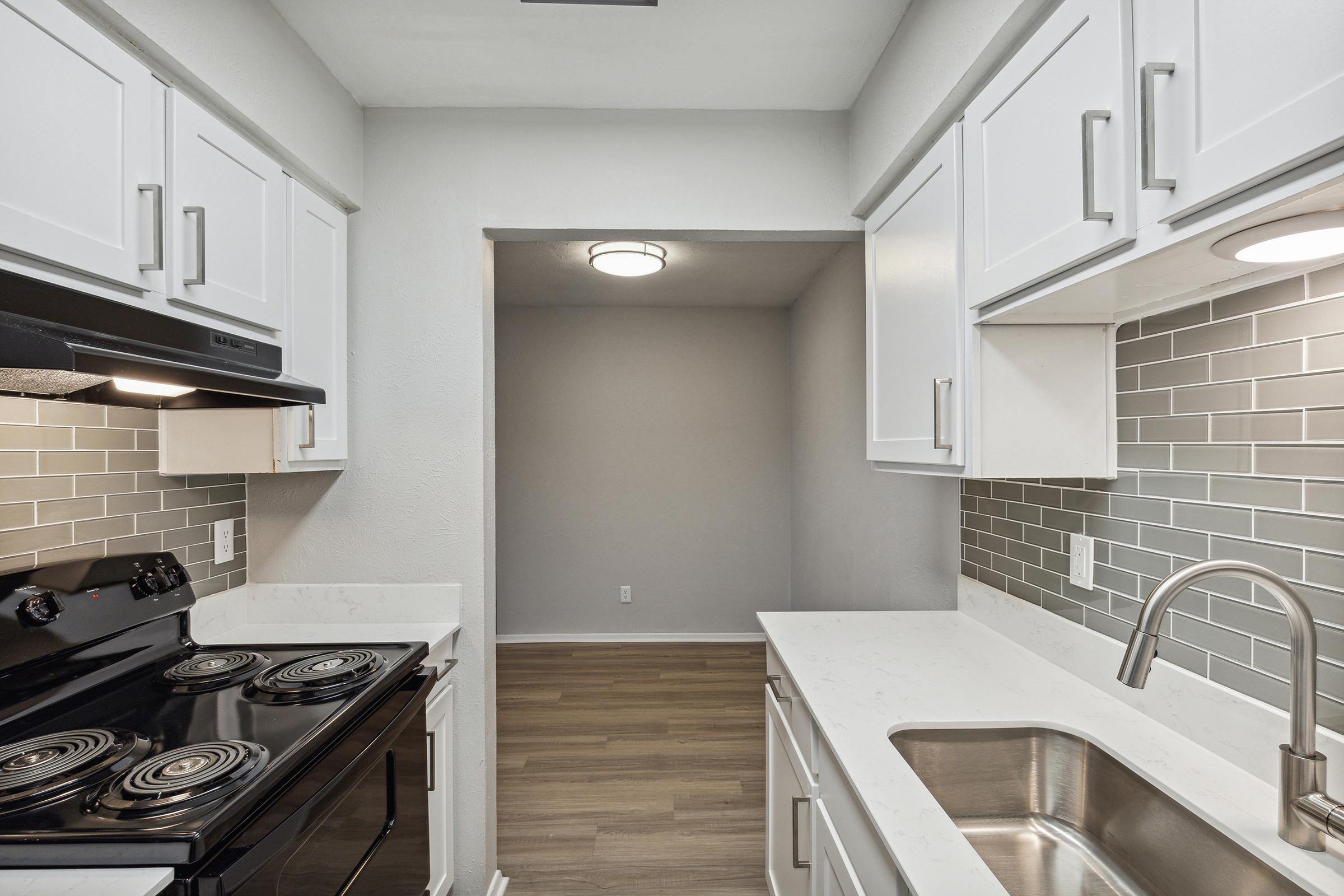 Modern kitchen with white cabinetry, a black stove, and a stainless steel sink. The backsplash features gray and white tiles. There is a small adjoining area visible through an open doorway, with neutral-colored walls and laminate flooring. Bright overhead lighting provides a warm ambiance.