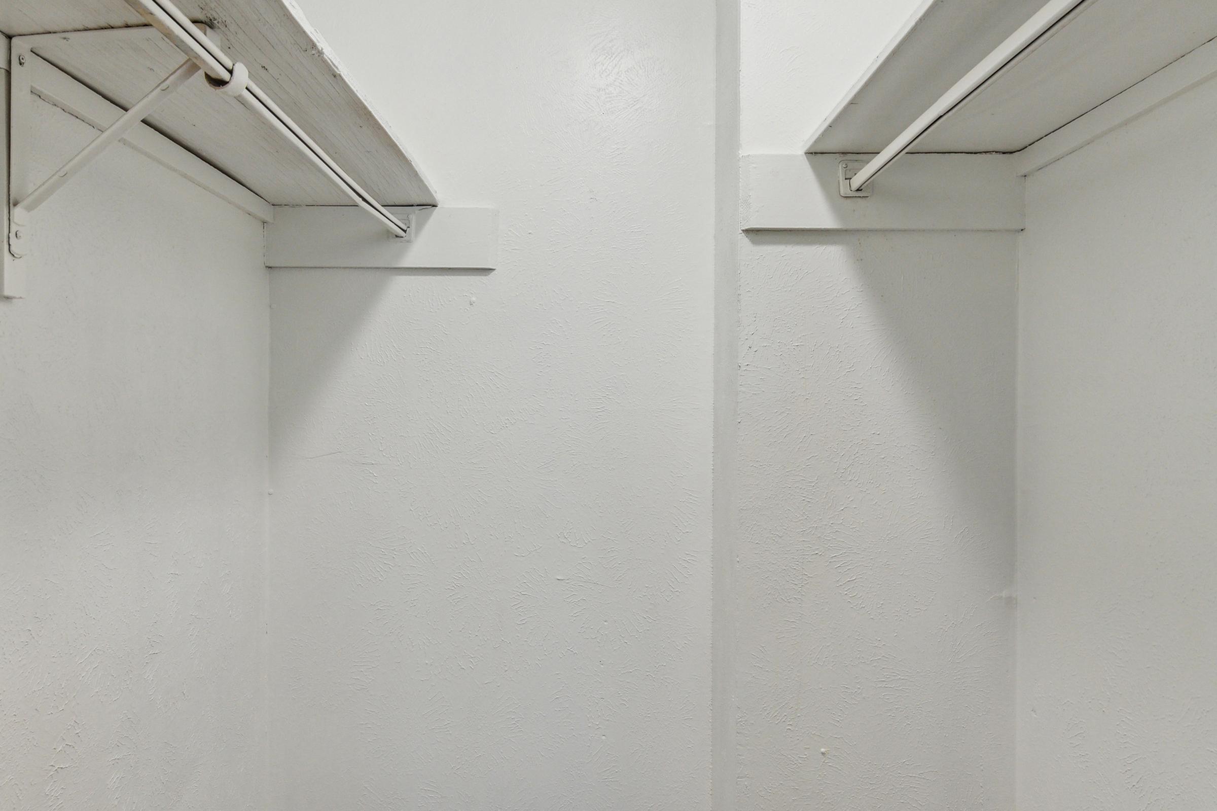 A simple, empty white closet space with walls that have a textured finish. There are two horizontal metal rods mounted on the walls for hanging clothes, with no visible items or decorations. The lighting is bright, emphasizing the clean and minimalist appearance of the space.