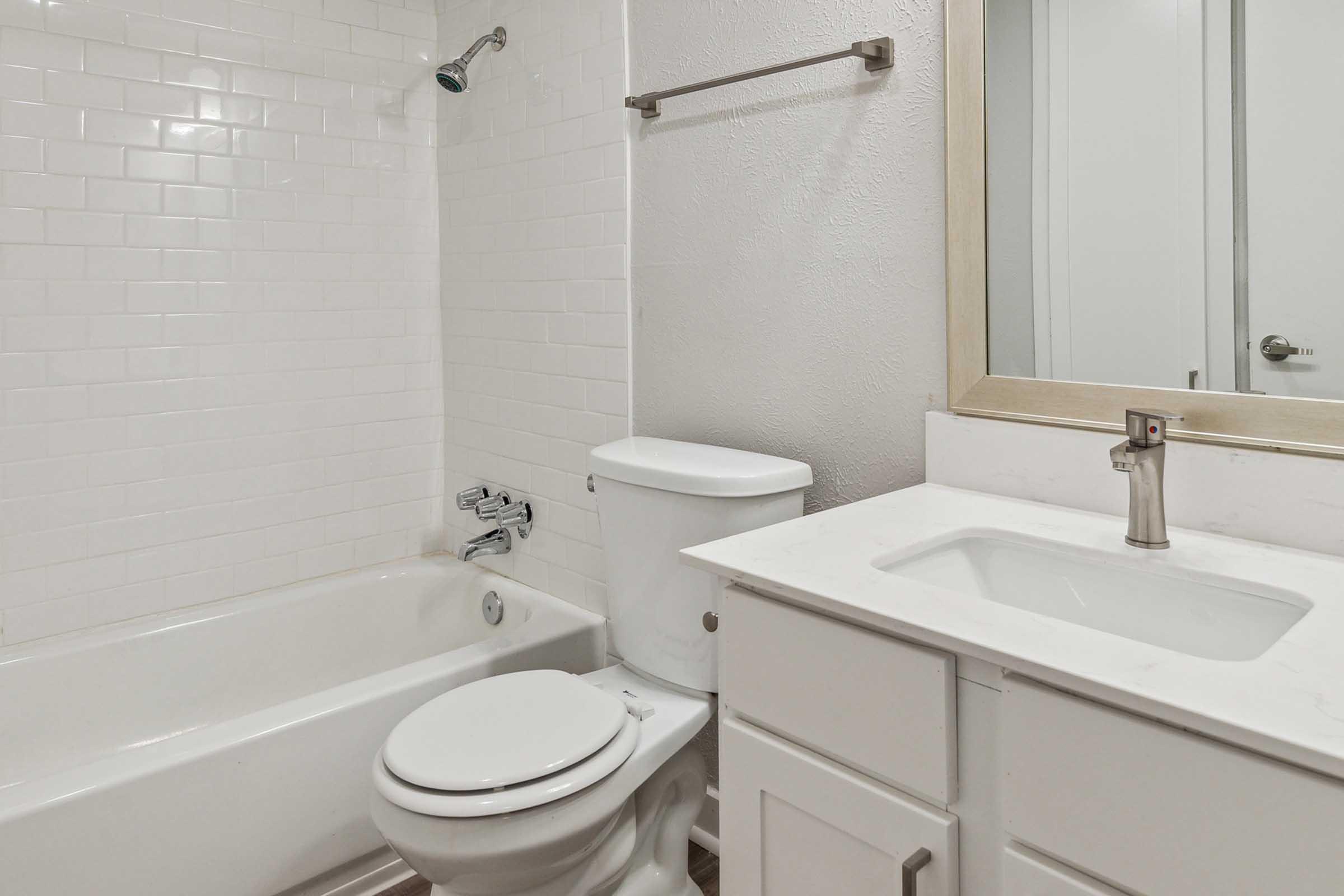 A clean and modern bathroom featuring a white tiled shower area with a bathtub, a white toilet, a white vanity with a single sink, and a mirror above the sink. The overall design is bright and minimalist, with neutral colors and ample natural light.