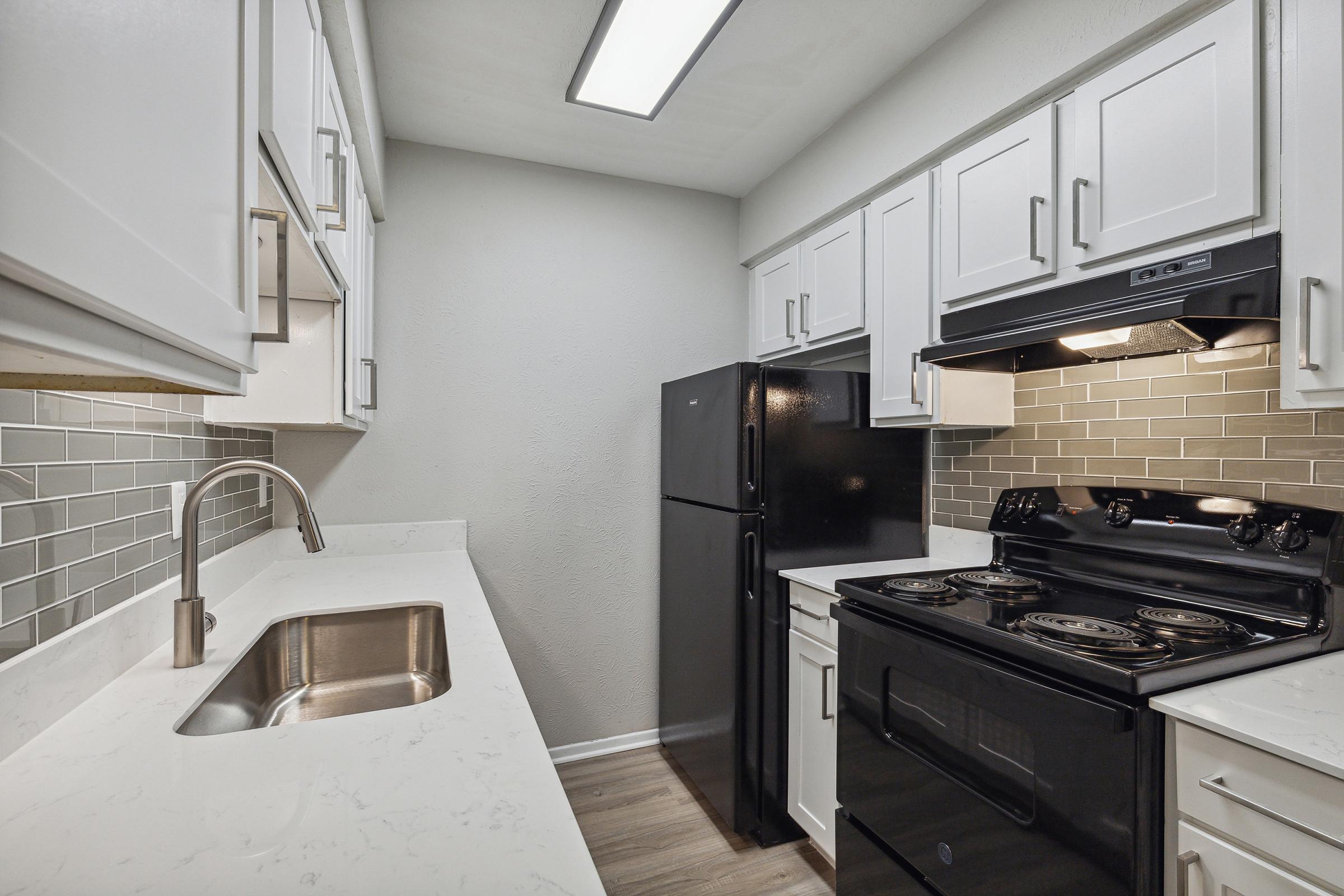 A modern kitchen featuring white cabinetry, a black refrigerator, and a black stove with an overhead microwave. The countertops are light-colored with a stainless steel sink, and the backsplash consists of gray tiles. The space is well-lit with a ceiling light.