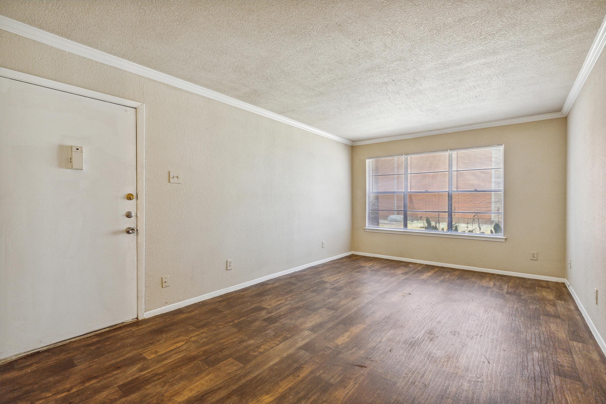Empty room with light-colored walls and a wooden floor. There is a white door on the left side and a large window on the right side allowing natural light to enter. The window frames an outdoor view, while the room is free of furniture, creating an open and spacious feel.