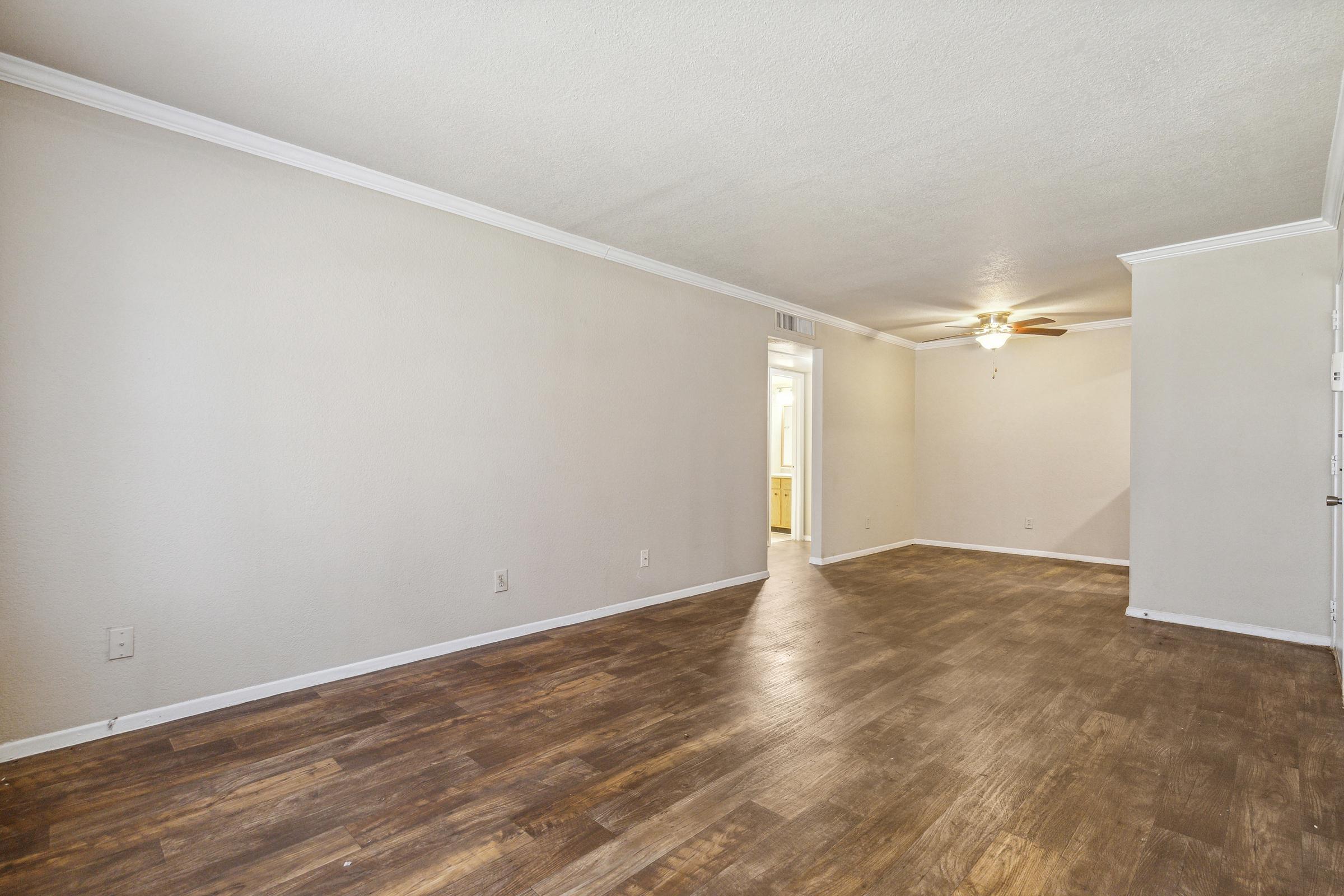 Spacious empty living room with light-colored walls, hardwood flooring, and a ceiling fan. A doorway leads to a kitchen area in the background. Natural light enters from the left side of the room.