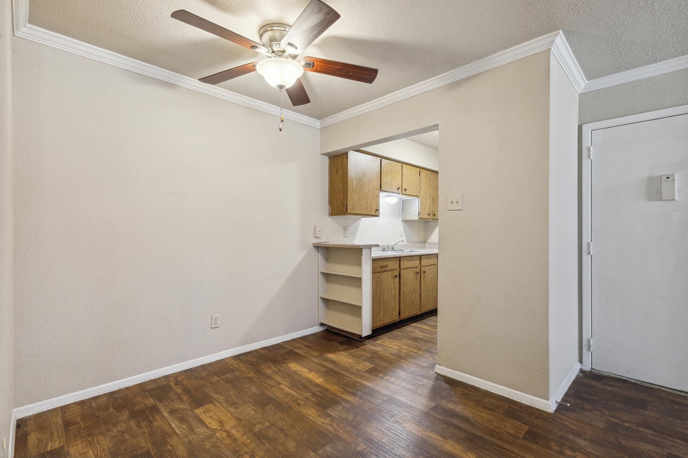 A small living area featuring a ceiling fan, hardwood floors, and beige walls. A doorway leads to a kitchen with wooden cabinets and a light above a countertop. There's also a door on the right, possibly leading to an entrance or hallway. The space is well-lit and has a cozy, inviting feel.