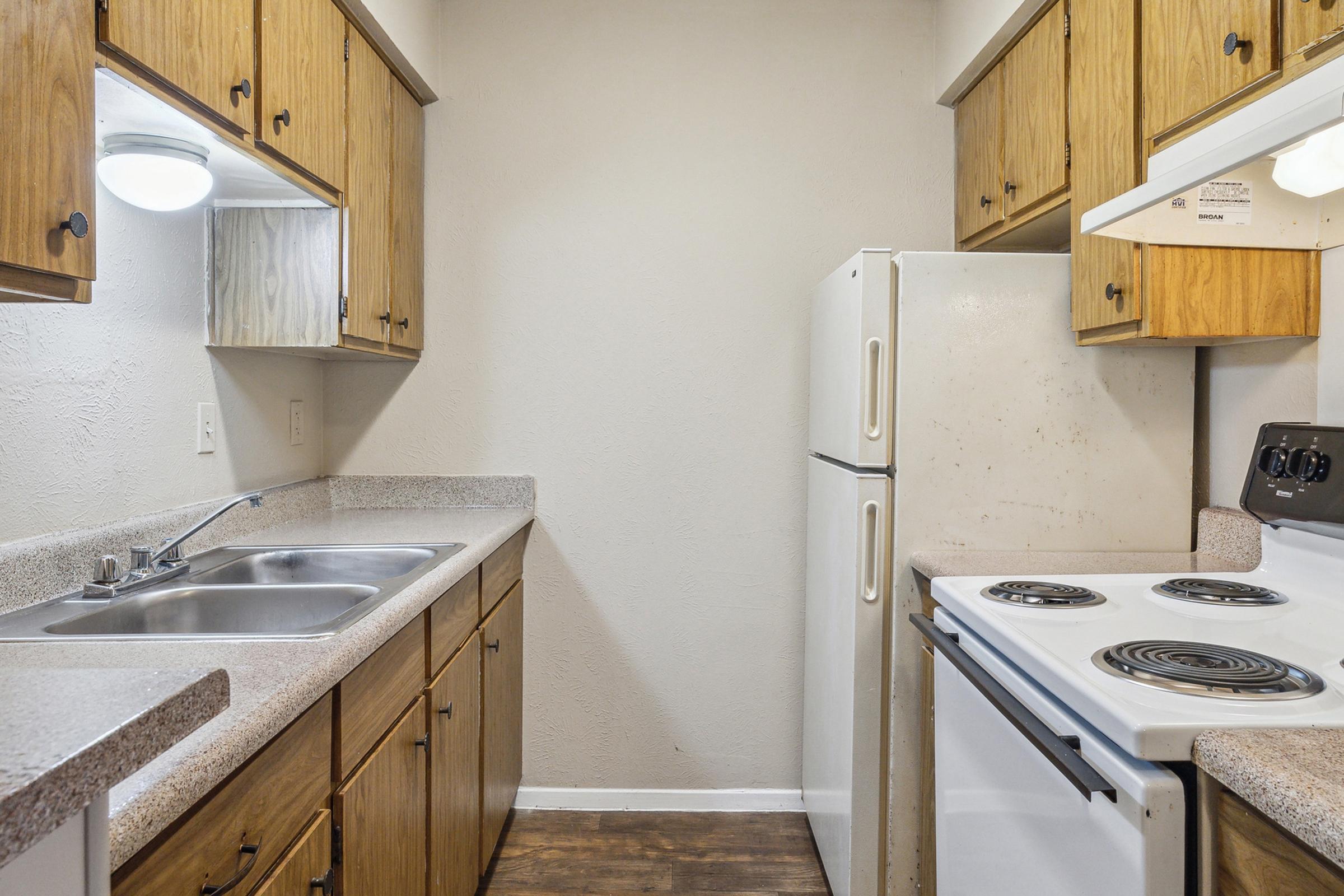A small kitchen featuring wooden cabinets, a double sink, a white refrigerator, and a white electric range with a black stove top. The walls are a light color, and the floor is a wood-like laminate. Natural light comes from a ceiling fixture and a mirror reflecting the space.