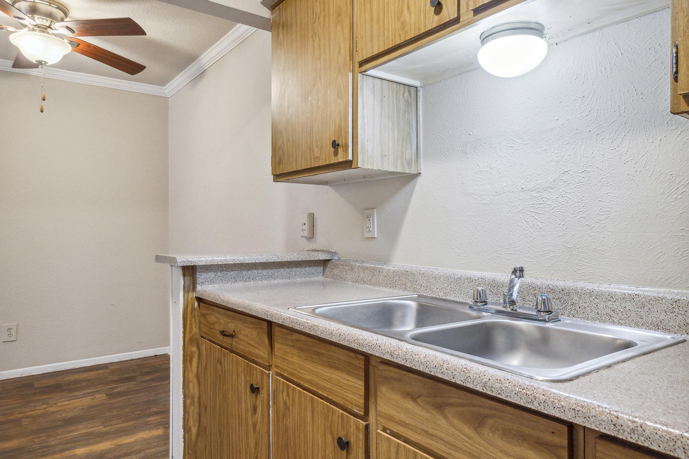 A kitchen with wooden cabinets, double sink, and stone countertop. A ceiling fan is visible in the background, along with a light fixture above the sink. The walls are painted a light color, and the flooring appears to be wooden. The space is tidy and well-lit, suggesting a functional kitchen area.