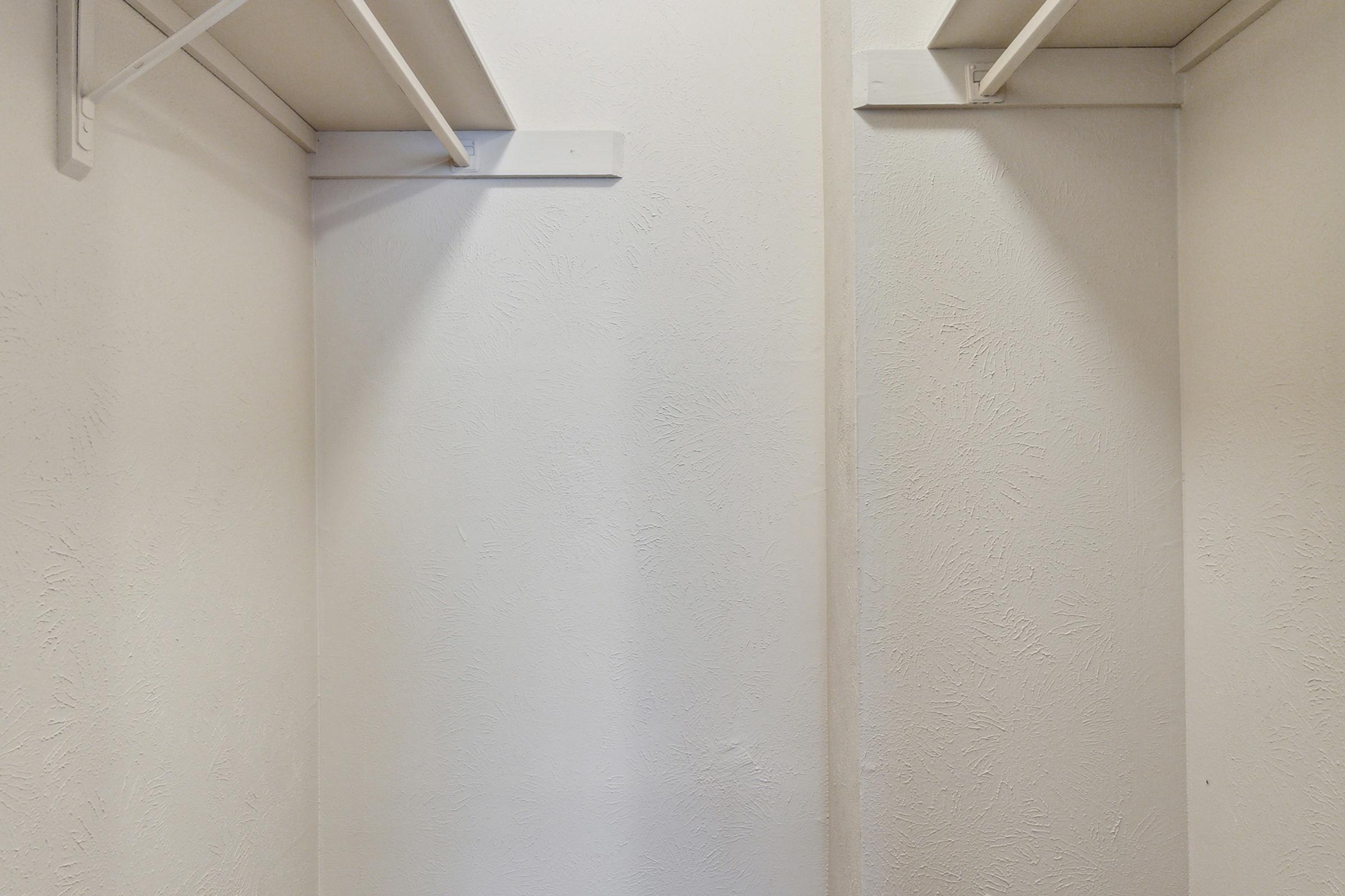 A simple, empty closet with white walls and a few hanging shelves. The space is well-lit, showcasing the plain texture of the walls and the organization of the shelving.