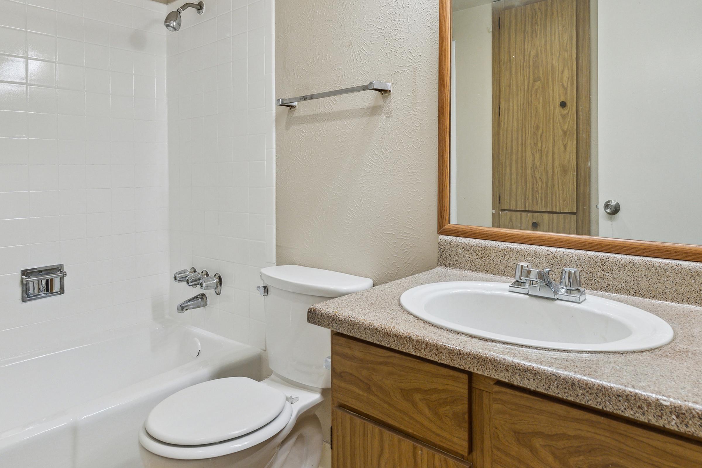 A clean, modern bathroom featuring a beige countertop with a sink, a wooden cabinet underneath, a white toilet, and a shower/tub combination with white tiles. A mirror is mounted above the sink, and a towel rack is visible on the wall. The space is well-lit and tidy.