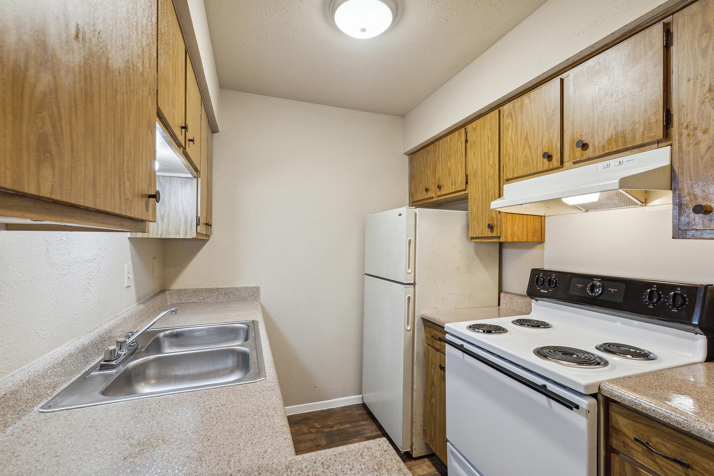 A small kitchen featuring wooden cabinetry, a white refrigerator, a black stove, and a ventilator. The countertop has a double sink, and the walls are painted a light color, creating a cozy and functional cooking space. Natural light fills the room from a ceiling light fixture.