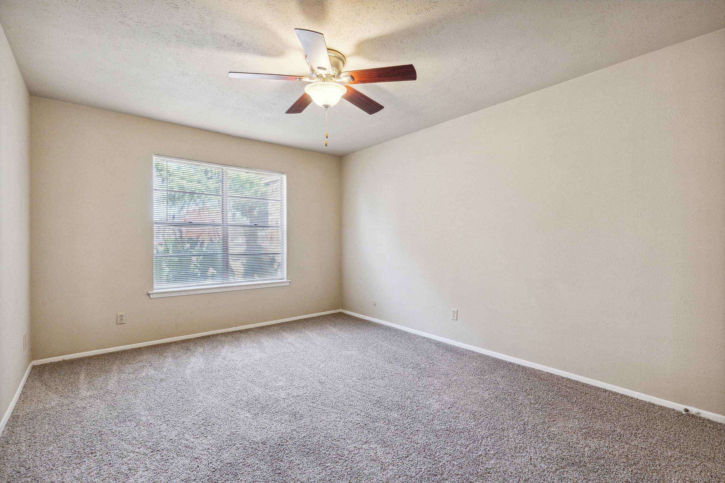 A spacious, empty room with light beige walls and carpeted flooring. A ceiling fan with wooden blades hangs from the ceiling. The room features a large window with blinds, allowing natural light to illuminate the space.