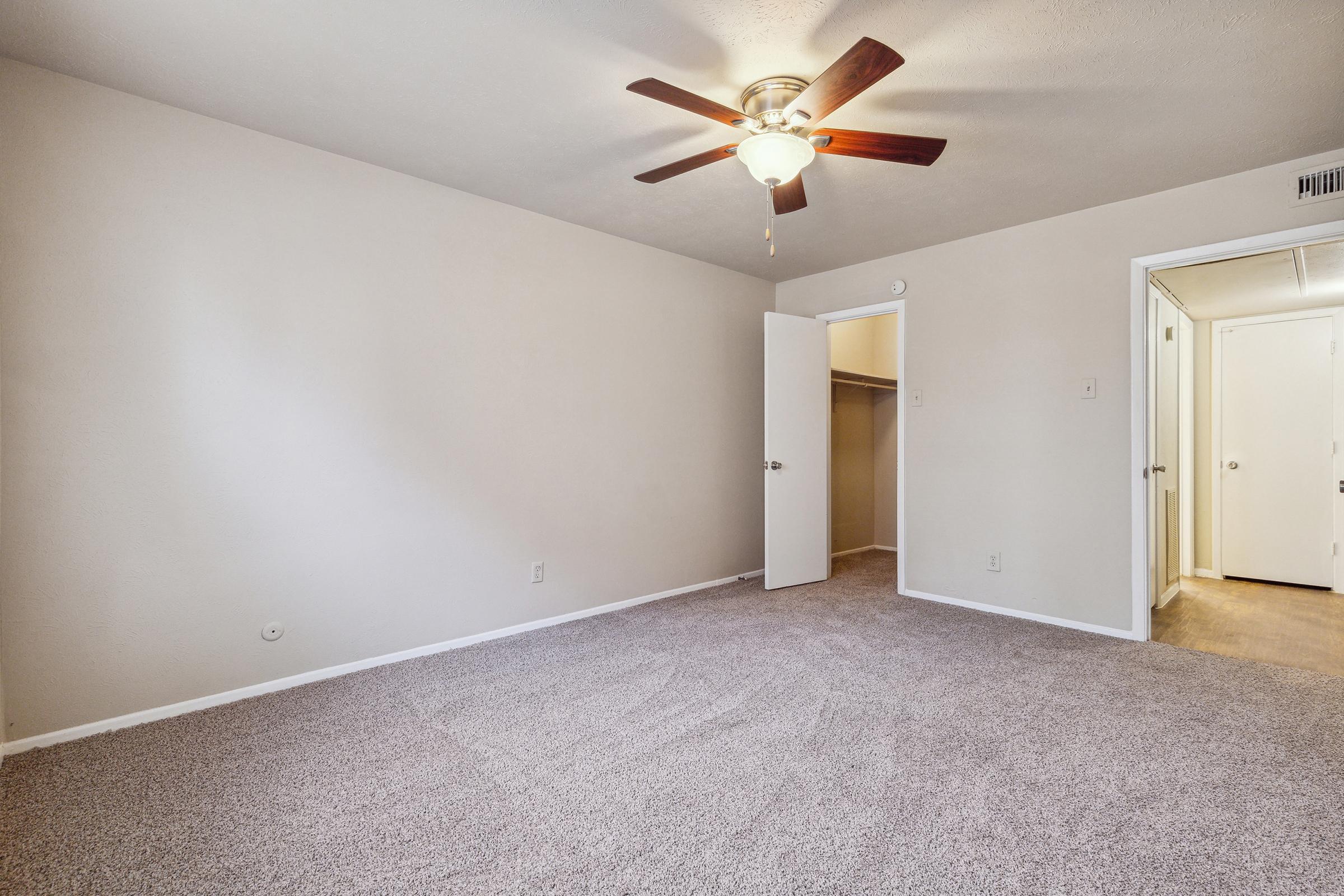 A well-lit, empty room with beige walls and carpet. It features a ceiling fan with wooden blades and an open closet with doors. There are two doors leading to adjacent spaces, emphasizing the room's spaciousness and simplicity. Ideal for a bedroom or office setup.