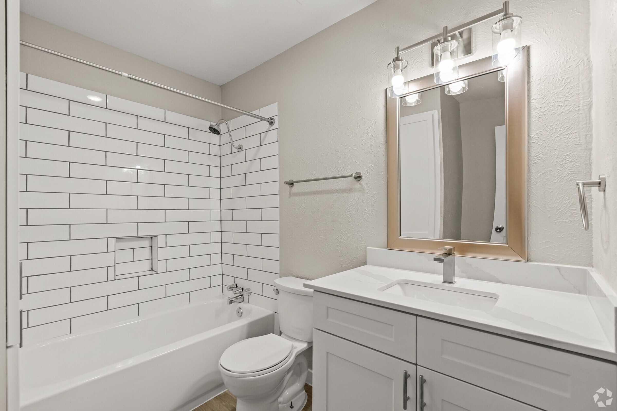 A modern bathroom featuring a white tiled shower with a bathtub, a stylish mirror above a sink with a sleek countertop, and light fixtures. The walls are painted in a neutral tone, and the floor has a wood-like appearance, creating a cozy and inviting space.