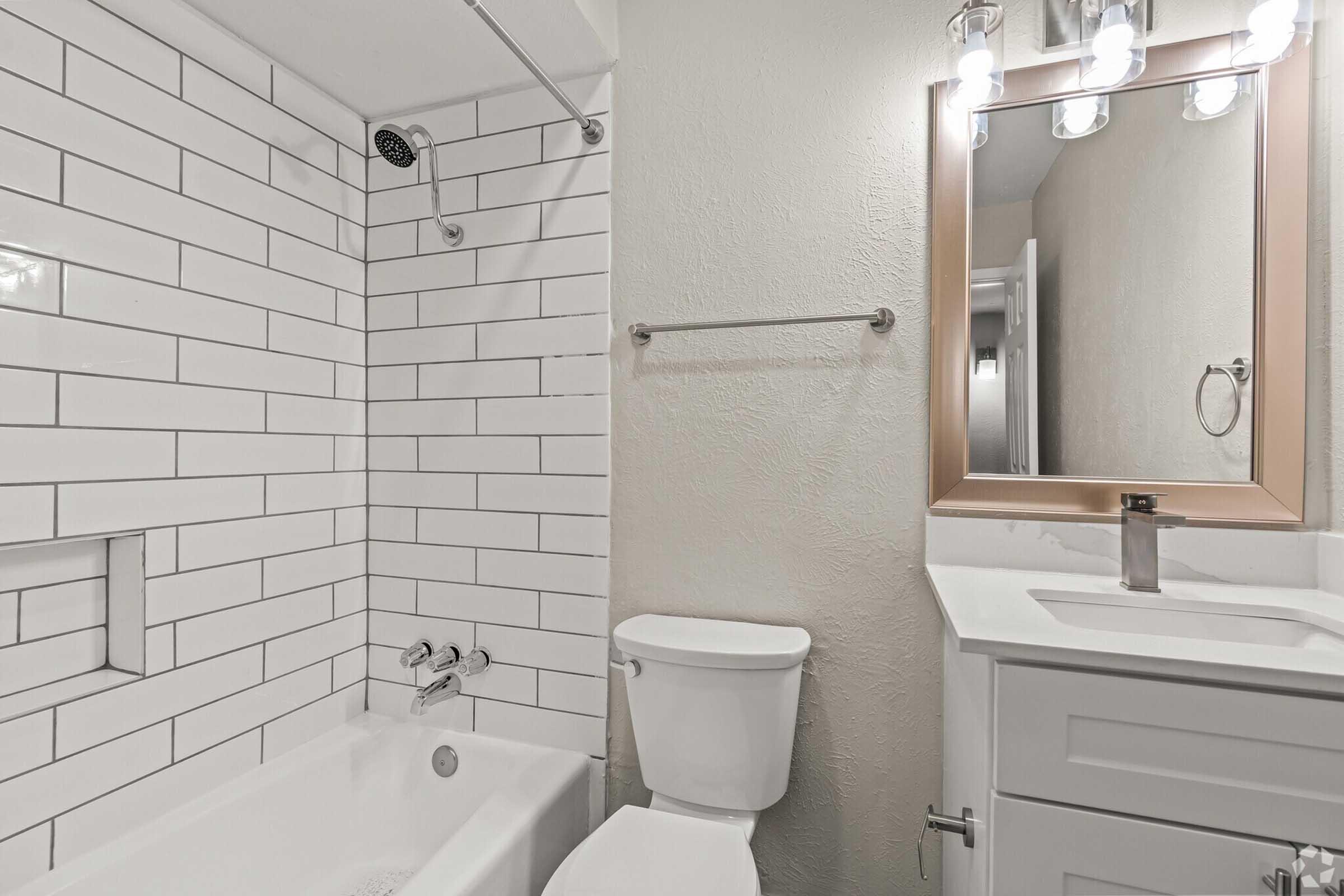 A modern bathroom featuring a white tiled shower and bathtub combination, a wall-mounted mirror with soft lighting, a sleek sink with a cabinet, and a toilet. The design is clean and minimalistic, showcasing neutral colors and contemporary fixtures.