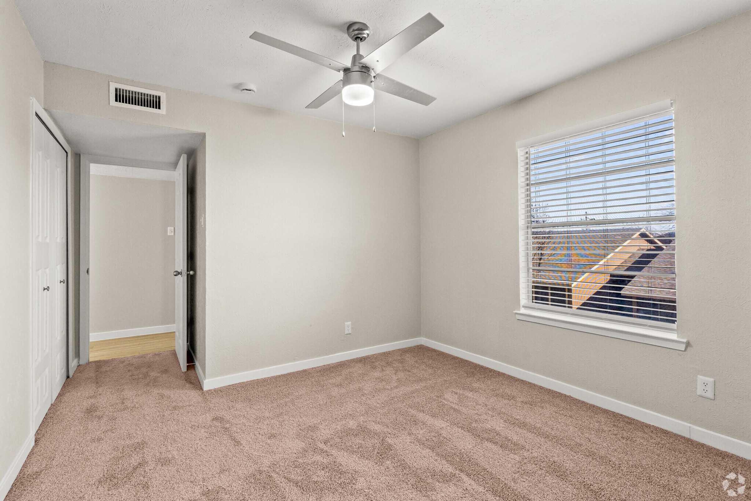 A vacant room with light-colored walls and plush beige carpet. A ceiling fan is mounted in the center, and there is a window with blinds on the right, allowing natural light to enter. A door is visible on the left, leading to another area of the space. The room has a clean, minimalistic design.