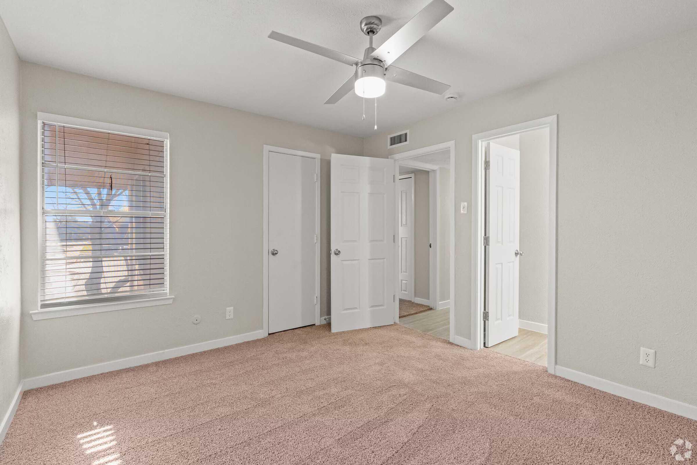 A well-lit, empty room featuring light beige carpet, a ceiling fan, and white walls. There are two doors leading to different areas, and a window with blinds allowing natural light to enter. The room has a clean and modern appearance, suitable for various uses.