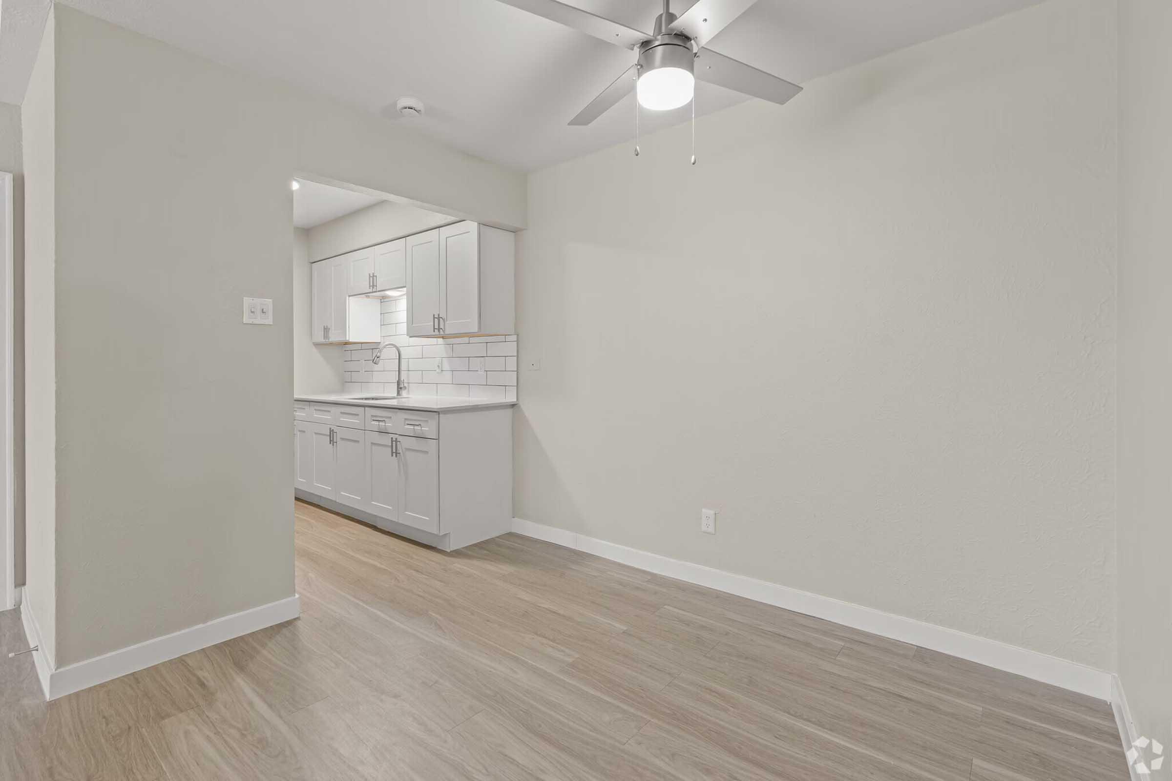 A bright, minimalist room with light gray walls and a wooden floor. A ceiling fan is visible, and an open doorway leads to a modern kitchen with white cabinets and a tiled backsplash. Natural light illuminates the space, creating a clean and airy atmosphere.