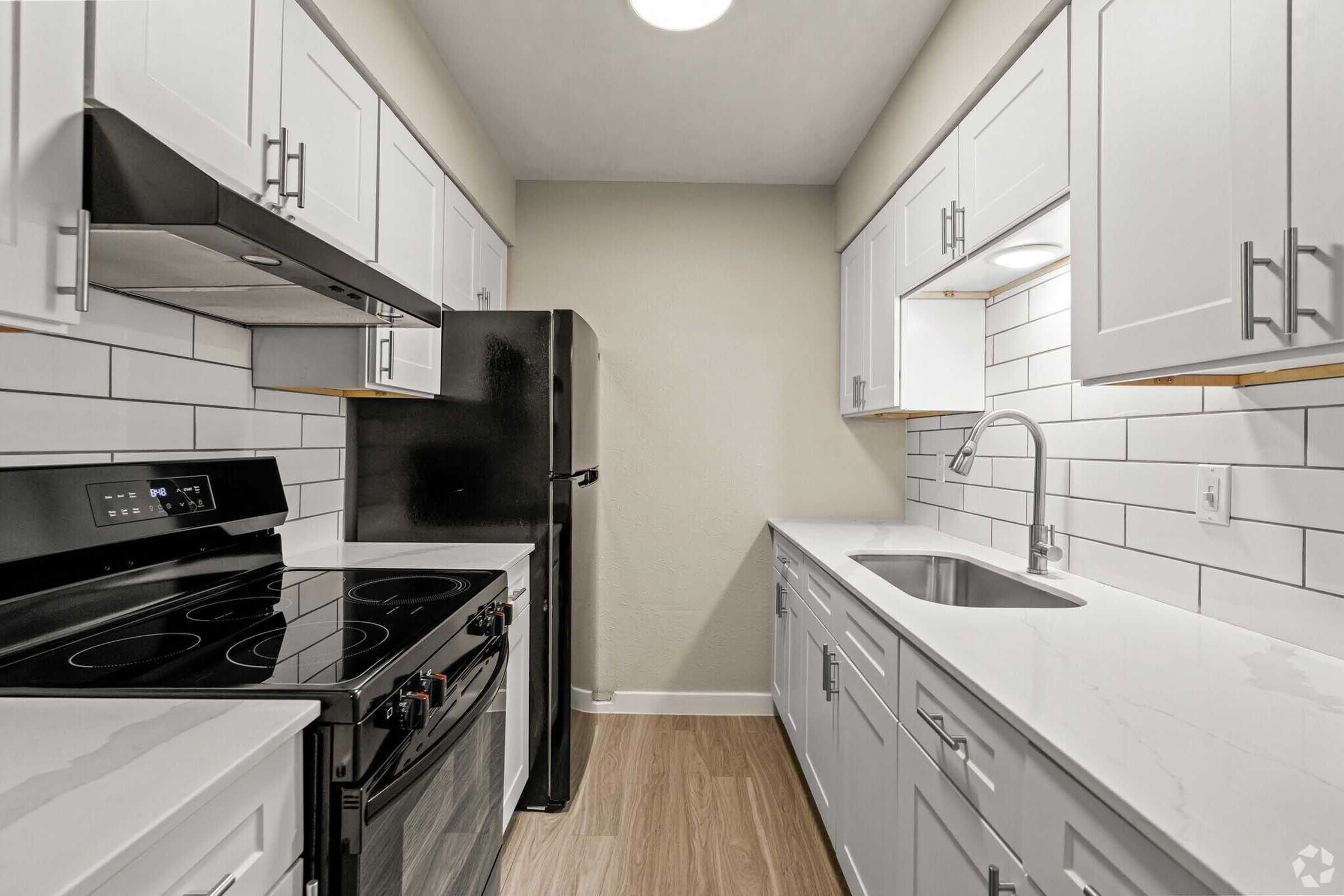 A modern kitchen featuring white cabinets, stainless steel appliances, and a sleek countertop. The layout includes a black refrigerator, an oven, a stove, and a sink. The backsplash consists of white tiles, creating a bright and clean aesthetic.