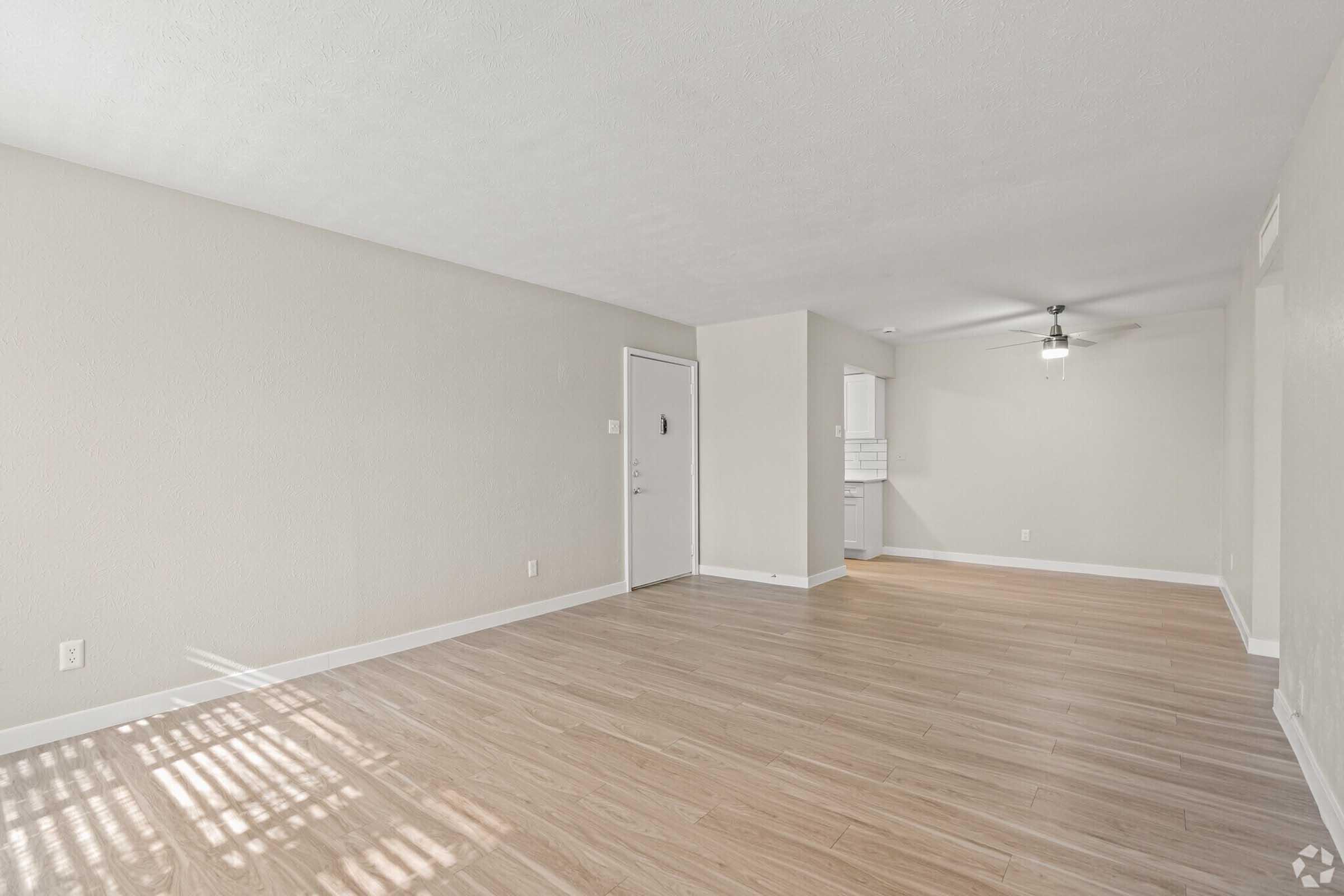 A spacious, empty living room with light-colored walls and hardwood floors. There's a front door on the left and a small kitchenette area visible in the background. Natural light streams in through a window, casting soft shadows on the floor. A ceiling fan is mounted in the corner.
