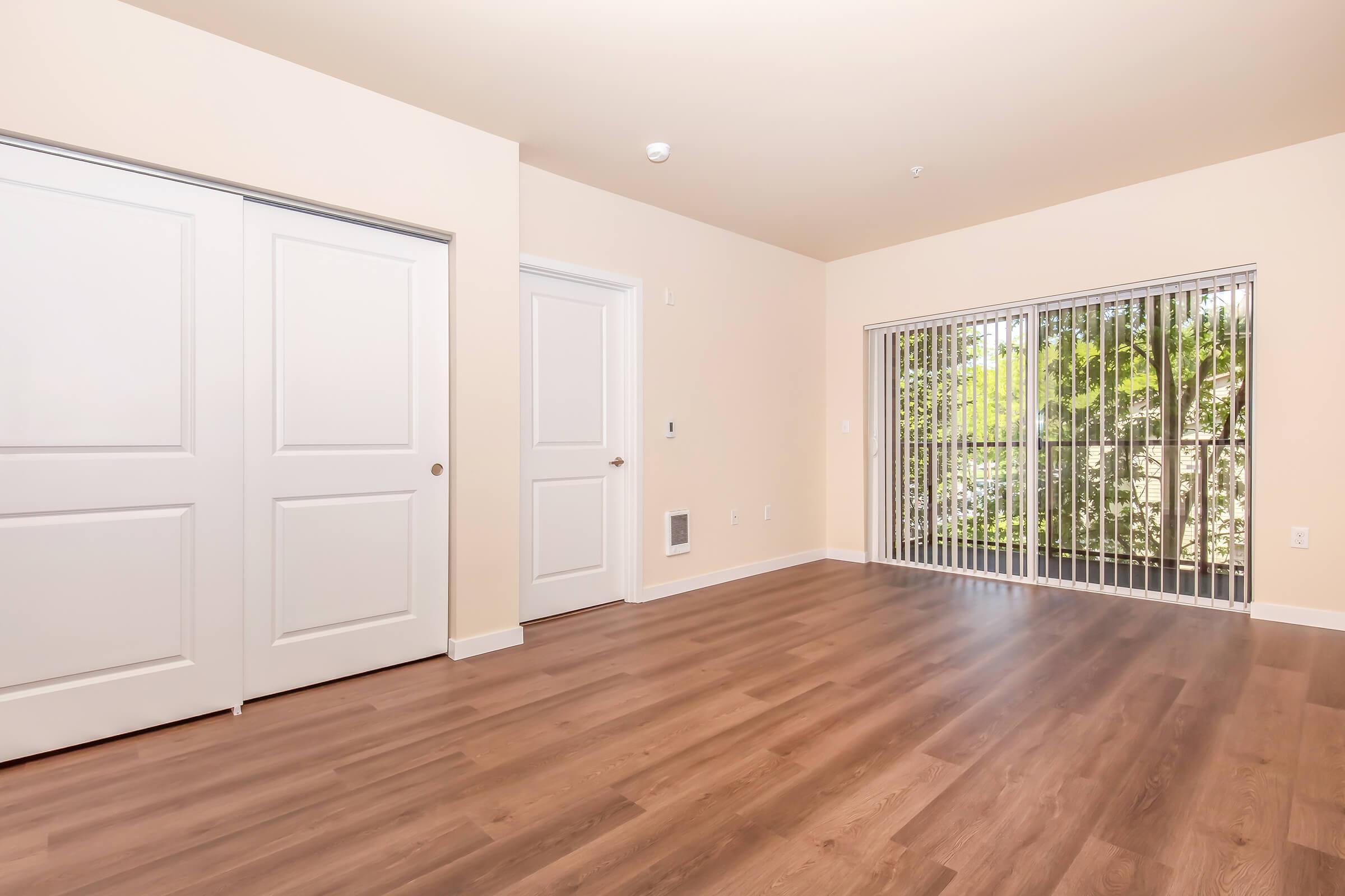 A spacious interior of a room featuring a wooden floor, two doors leading to closets, and a large window with vertical blinds revealing a view outside. The walls are a neutral color, creating a bright and open atmosphere.