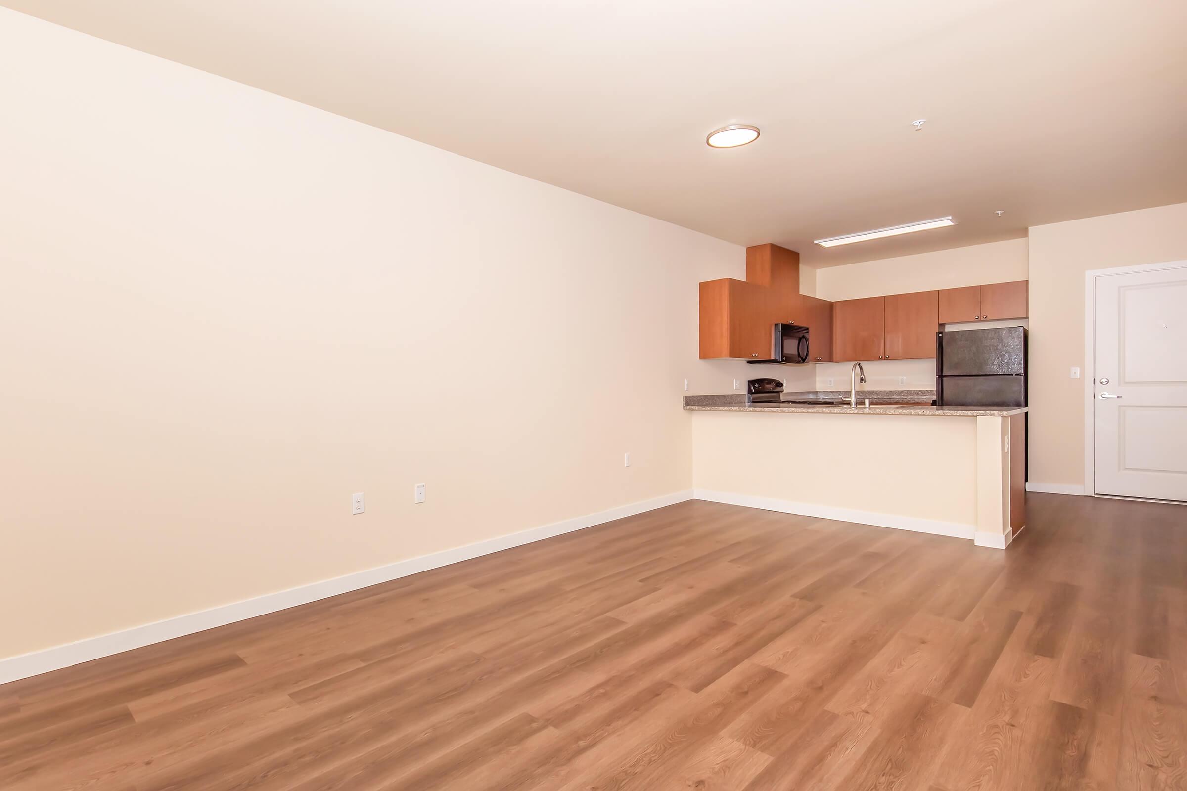 A spacious, empty apartment living area with light-colored walls and modern wood-style flooring. To the left, there's a kitchen area featuring wooden cabinets and a stainless steel refrigerator. The entrance door is visible on the right side, and the overall ambiance is bright and minimalist.