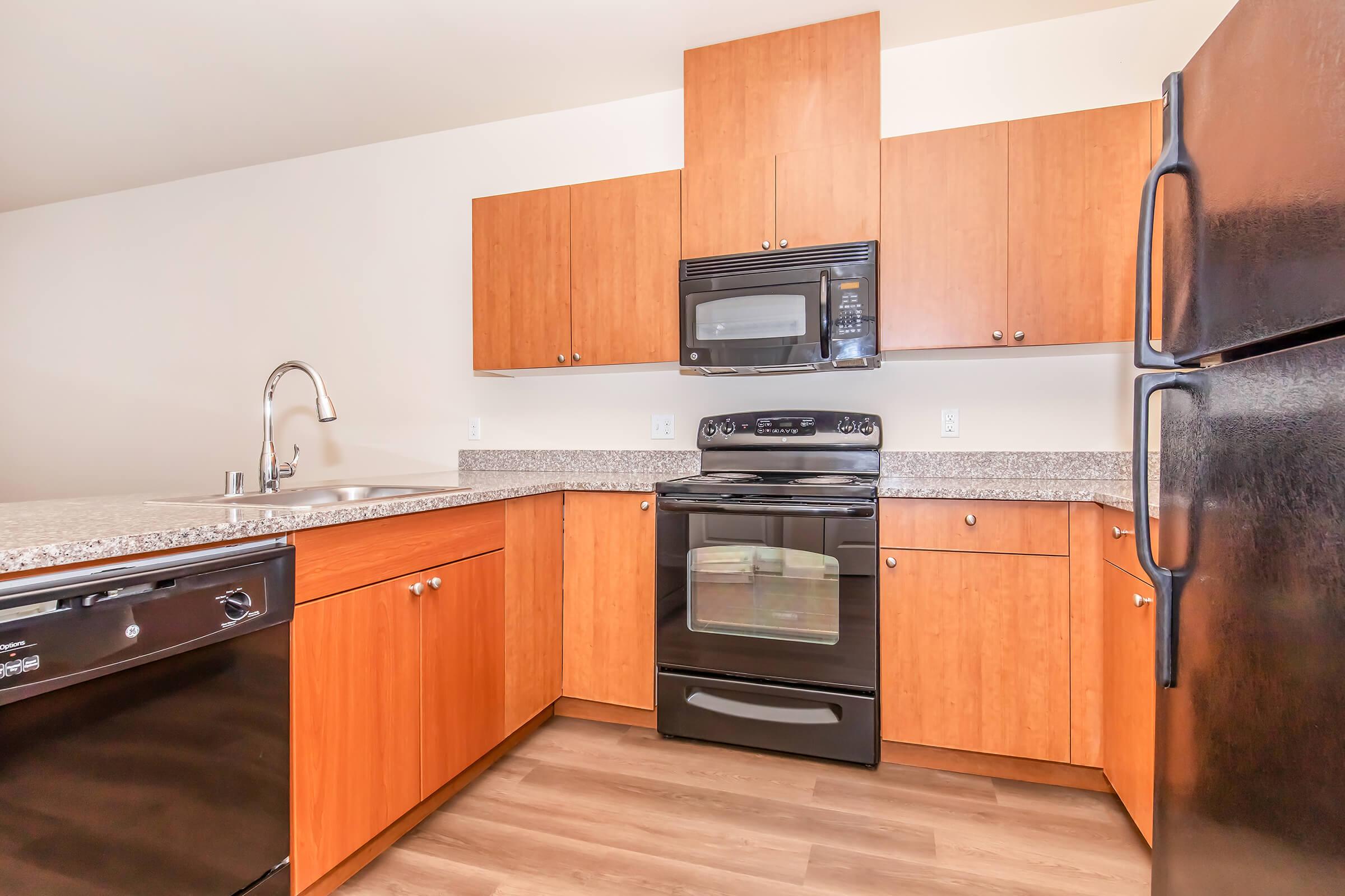 A modern kitchen featuring wooden cabinets, a granite countertop, a black microwave, and an oven. The kitchen includes a black dishwasher and a stainless steel sink with a faucet. The flooring is light-colored wood, and the walls are painted in a neutral shade.