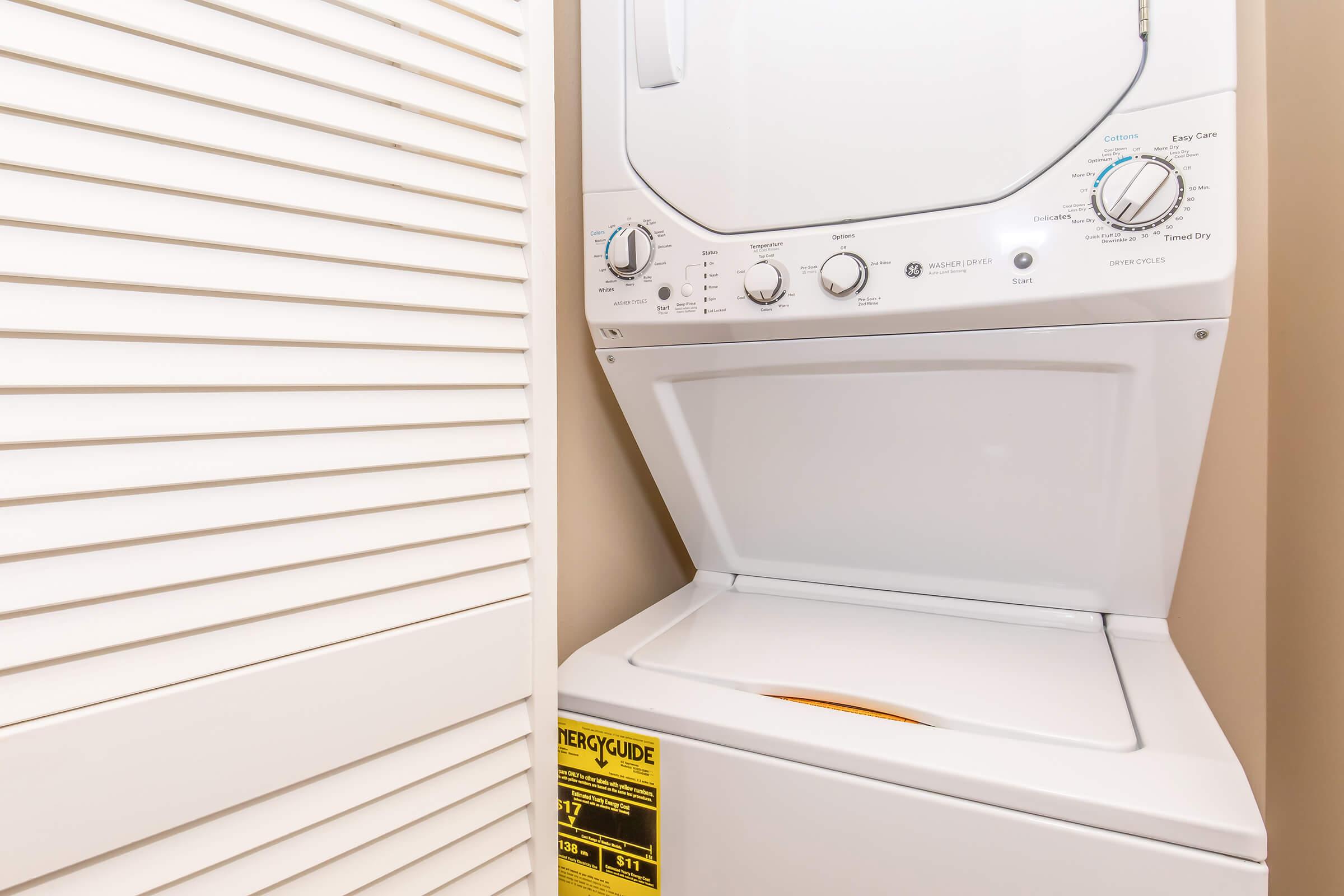 A stacked washer and dryer unit in a laundry space, positioned between two folding doors. The dryer features control knobs and a cycle selector, while the washing machine's top is partially visible. The appliance is white, with a bright yellow energy guide sticker on the front.