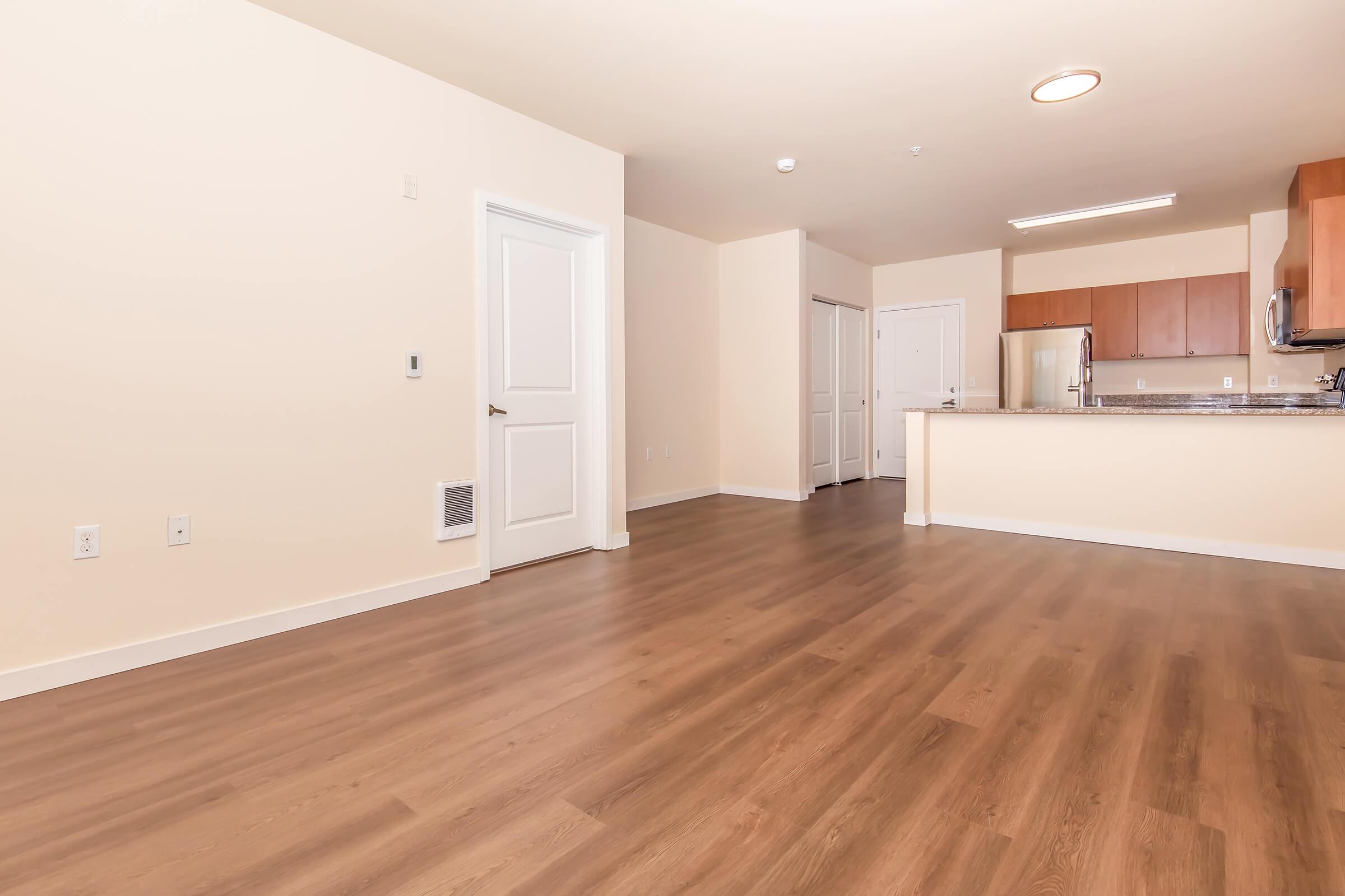 Spacious living area with hardwood-like flooring, featuring a doorway and a white kitchen counter with wooden cabinets in the background. Natural light fills the room from the open layout, showcasing a modern and inviting atmosphere. Neutral wall colors enhance the bright and airy feel.