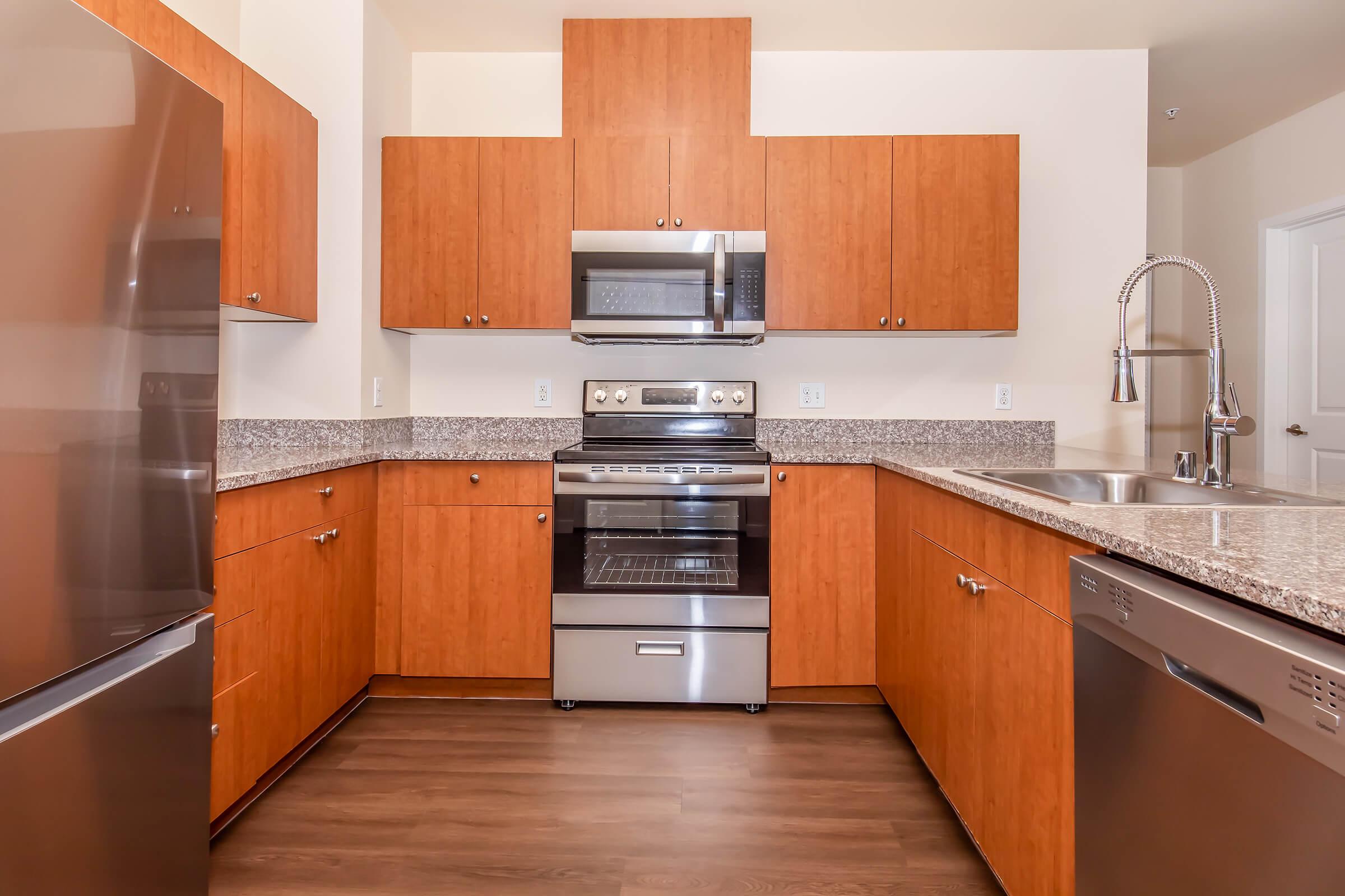 Modern kitchen featuring wood cabinetry, stainless steel appliances including a refrigerator, oven, and dishwasher, granite countertops, and hardwood flooring. Brightly lit with a spacious layout, showcasing an open design that emphasizes functionality and style.