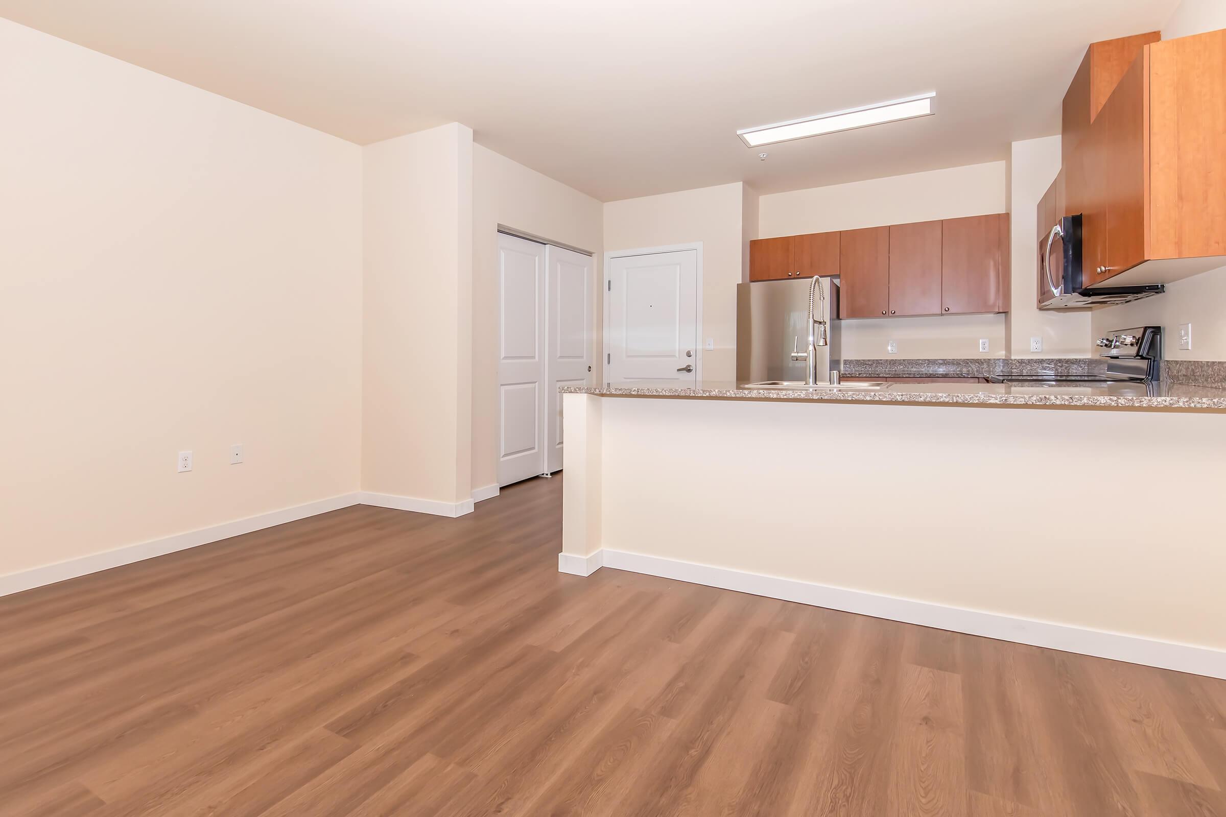 Modern, open-concept kitchen and living area featuring wood flooring, wooden cabinets, and stainless steel appliances. A doorway leads to another room, with a light and neutral color palette creating a spacious, airy feel.