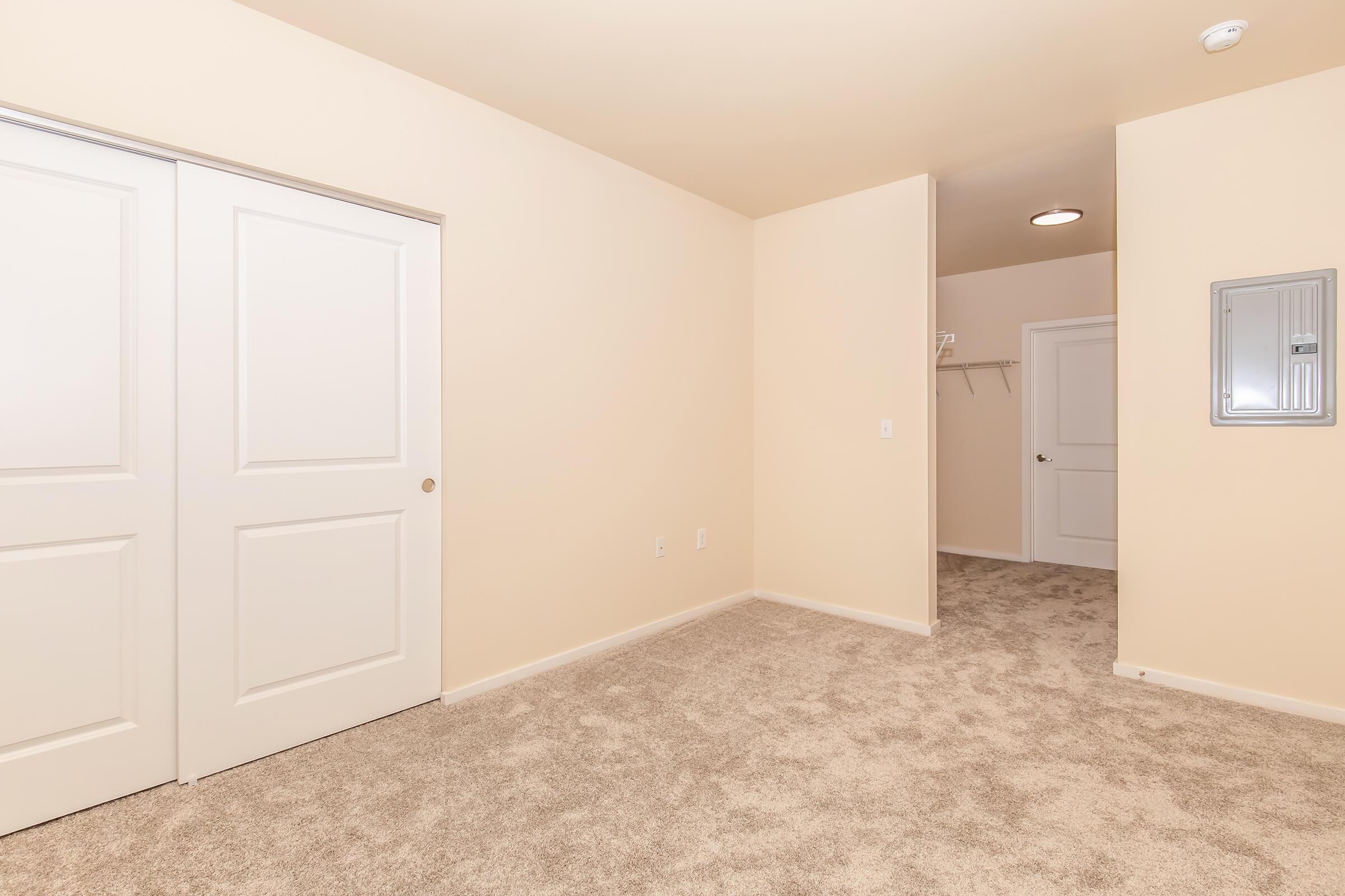 A spacious, empty room featuring beige walls, a carpeted floor, and two closed white doors on the left. There is a small entryway to the right, leading to another room, with a coat rack on the wall and an electrical panel mounted nearby. The overall ambiance is bright and uncluttered.